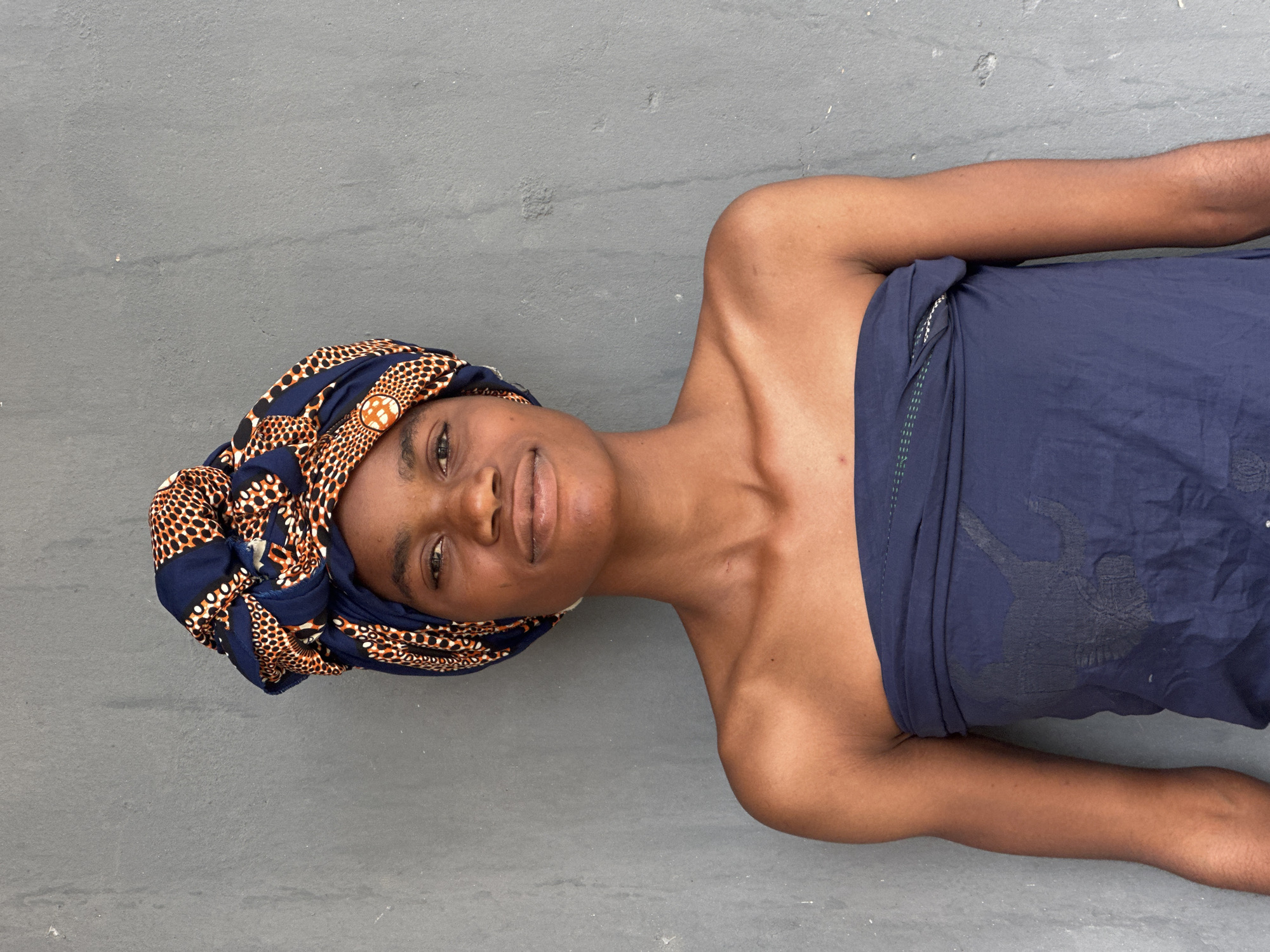 A young Black woman is standing against a gray wall in Badagry, Nigeria. She is wearing a dark blue strapless garment and a colorful headwrap with a pattern of navy blue, orange, and white circles. Her shoulders and upper chest are visible. The wall behind her is textured and appears to be made of concrete. The lighting suggests it is daytime.