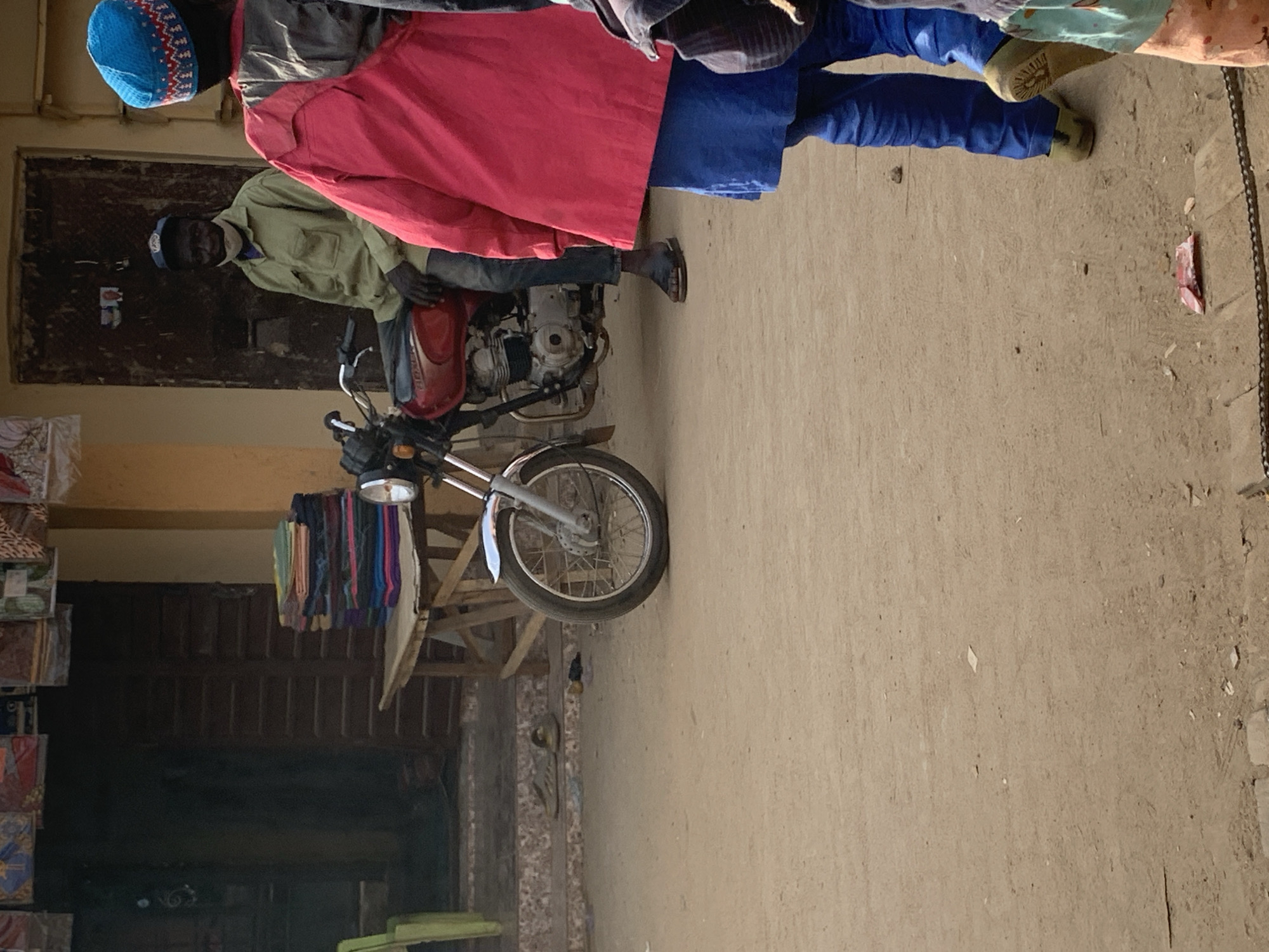 The scene is in Kafanchan B, Nigeria. A motorcycle is parked on a dirt or concrete surface. A man wearing a light green shirt, blue pants, and dark shoes is sitting on the motorcycle. Behind him, another person wearing a blue cap, grey jacket, red shirt, and blue pants stands beside the motorcycle. In the background is a building with a closed door and a vendor's display with textiles. A wooden structure is also visible near the textile display. The ground is dusty with some debris scattered around.