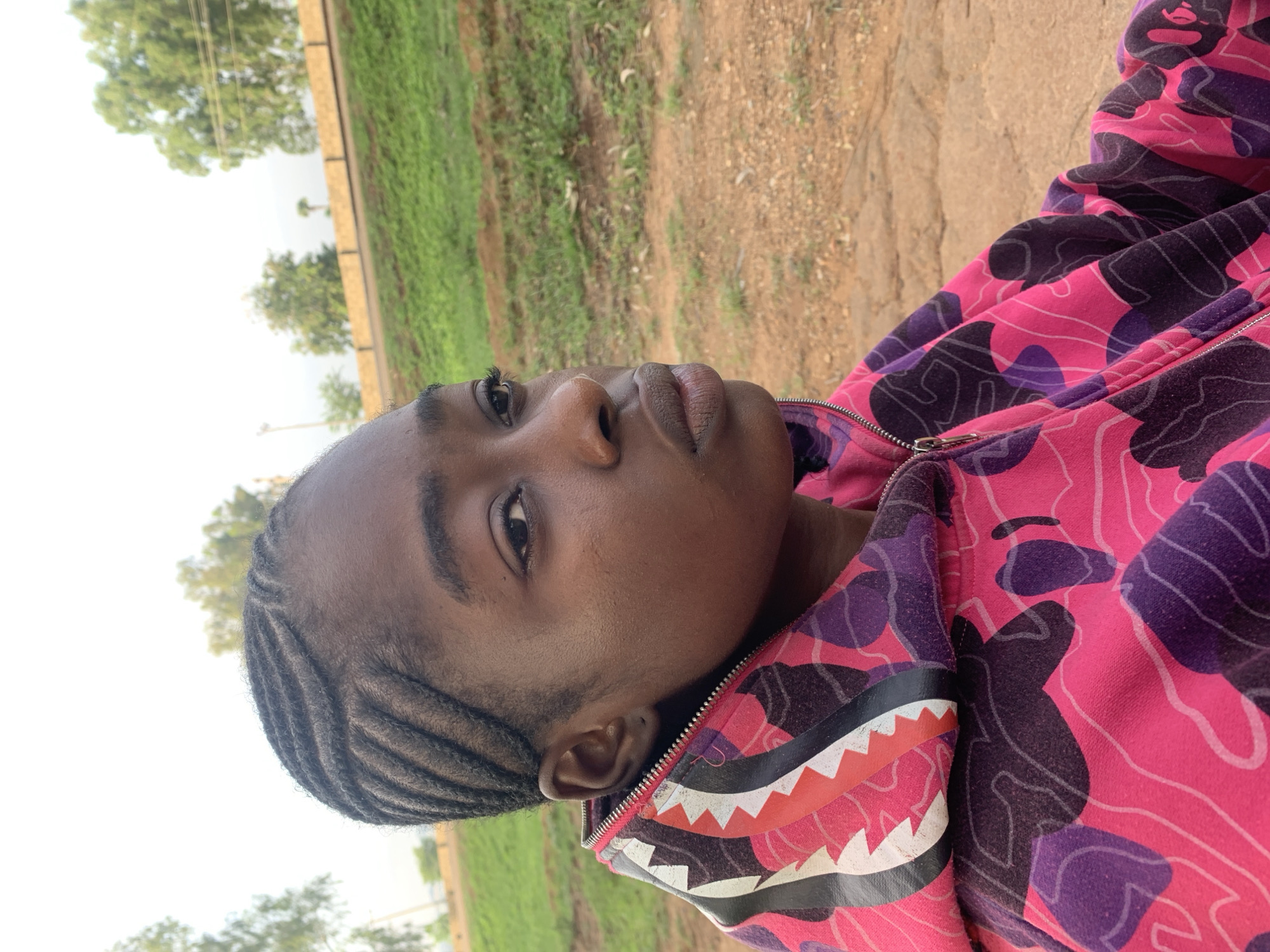 The image shows a close-up portrait of a young individual, likely female, with dark skin and hair styled in cornrows. The photograph is rotated 90 degrees, presenting the subject as if looking upwards and to the left. She wears a distinctive hooded jacket featuring a vibrant pink and purple camouflage pattern. A prominent design resembling white, black, and orange shark teeth is visible along the edge of the hood or collar, near the jacket's zipper.
The background depicts an outdoor scene under bright daylight. A bright, light-colored sky occupies the left side, while green vegetation, possibly grass and trees, is visible on the right. A dirt or gravel path runs along the right side of the frame. The lighting is bright and diffuse, suggesting an overcast day or indirect sunlight. The subject's expression is neutral and observant. There is no visible text in the image. The setting appears to be a rural or natural environment, consistent with Unguwar Saya, Nigeria.