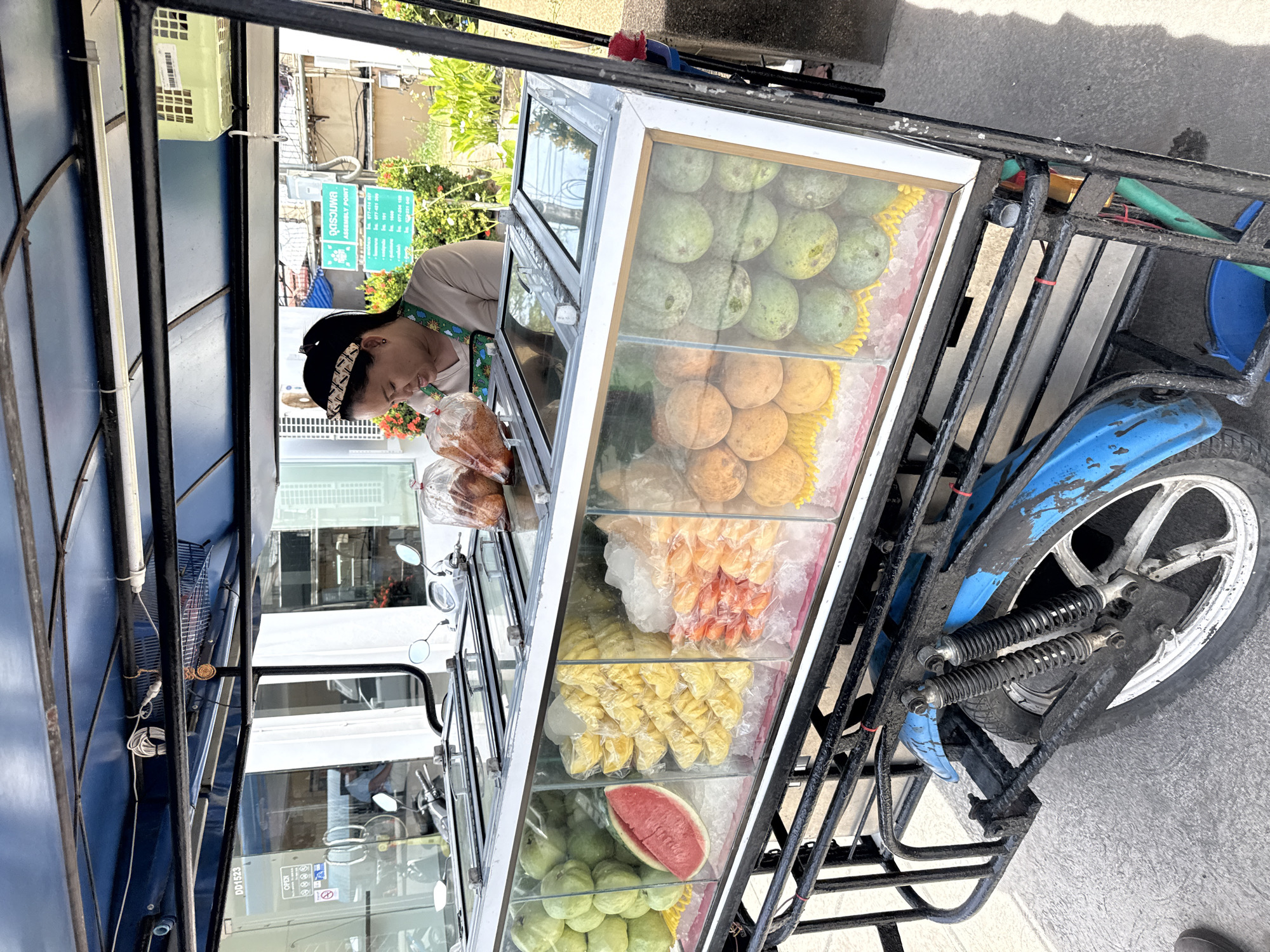 An outdoor fruit stand in Ko Samui District, Thailand, features a vendor serving customers from a refrigerated display cart. The cart is divided into compartments showcasing various fruits, including guavas, watermelon, pineapple, and santol. The vendor, wearing an apron and a headband, is seen preparing a customer's order. In the background, a building with signage and plants suggests a commercial area. The scene is illuminated by natural daylight.