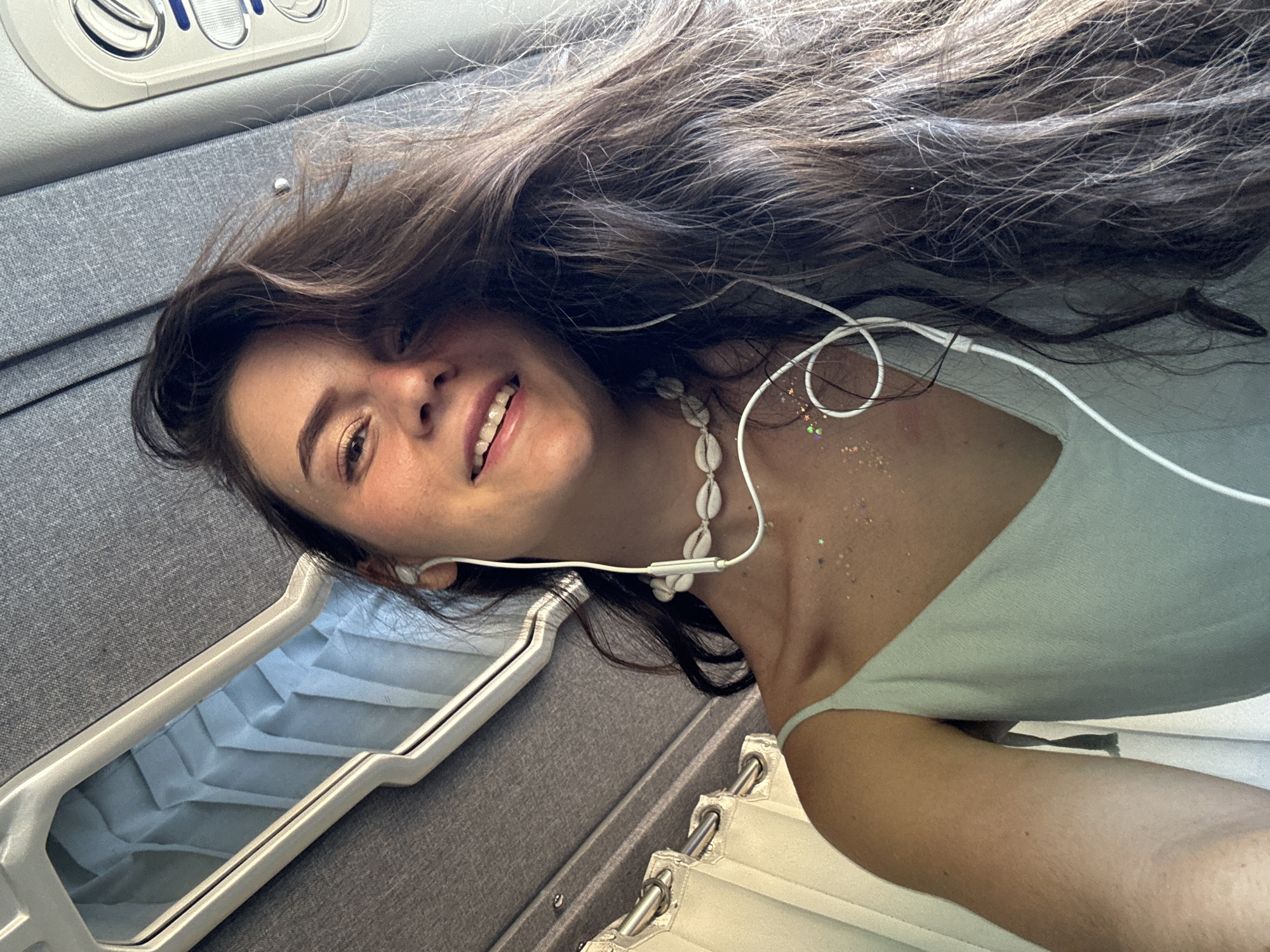 A young woman with long brown hair, fair skin, and a shell necklace is lying down. She is smiling and wearing earphones. Her hair is spread out across a grey, textured surface, possibly fabric. Her shoulder and chest are visible, adorned with small, iridescent sparkles. She is wearing a light green garment. To the left is an open, rectangular compartment with corrugated paper inside. The location is inferred to be Phu Thuy Ward, Vietnam.