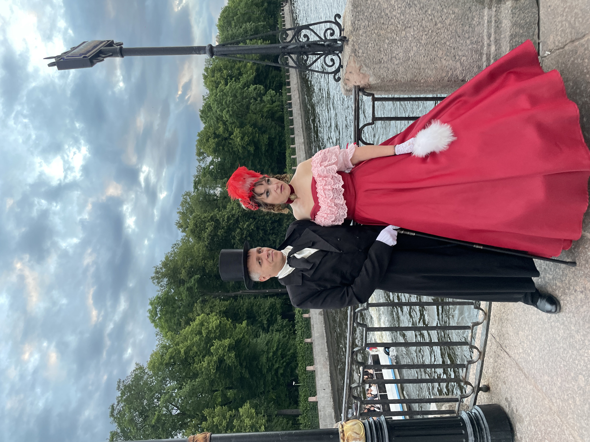Two individuals dressed in 19th-century attire stand on a stone bridge over a canal in Saint Petersburg, Russia. The man, on the left, wears a black tailcoat, white cravat, and a black top hat, holding a cane. The woman, on the right, is adorned in a vibrant red off-the-shoulder gown with pink lace trim and a red feathered headdress, carrying a white feather fan. They are posed with a body of water and a tree-lined embankment in the background. A boat with passengers is visible on the canal. The sky above is overcast with dramatic clouds.