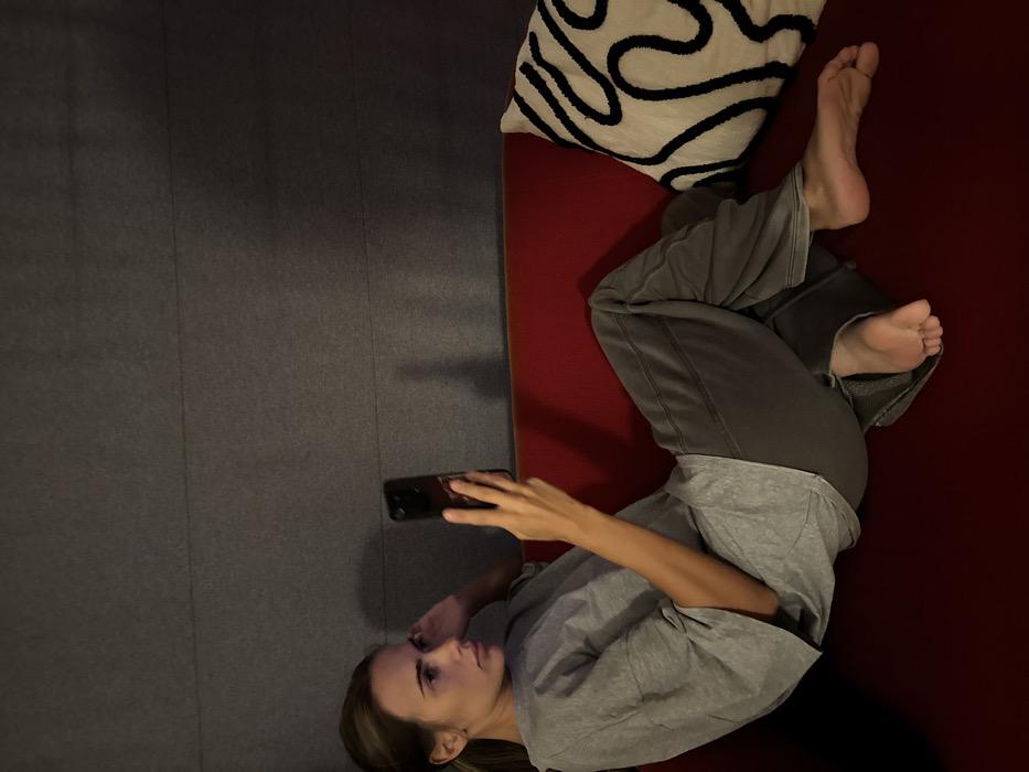 A female individual is observed reclining on a red upholstered sofa in an indoor setting in the city of Warsaw, Poland. She is positioned on her left side, facing left, with her left hand resting near her head. The subject wears a loose grey t-shirt and dark grey casual trousers, and is barefoot. She holds a dark-colored smartphone vertically with her right hand, directing her gaze towards its screen. Her legs are bent, with her feet visible. To the right of the individual, on the sofa, rests a decorative pillow featuring a white background with black abstract curvilinear patterns. The background wall is dark grey, appearing to be paneled horizontally. The scene is illuminated by dim, ambient light.