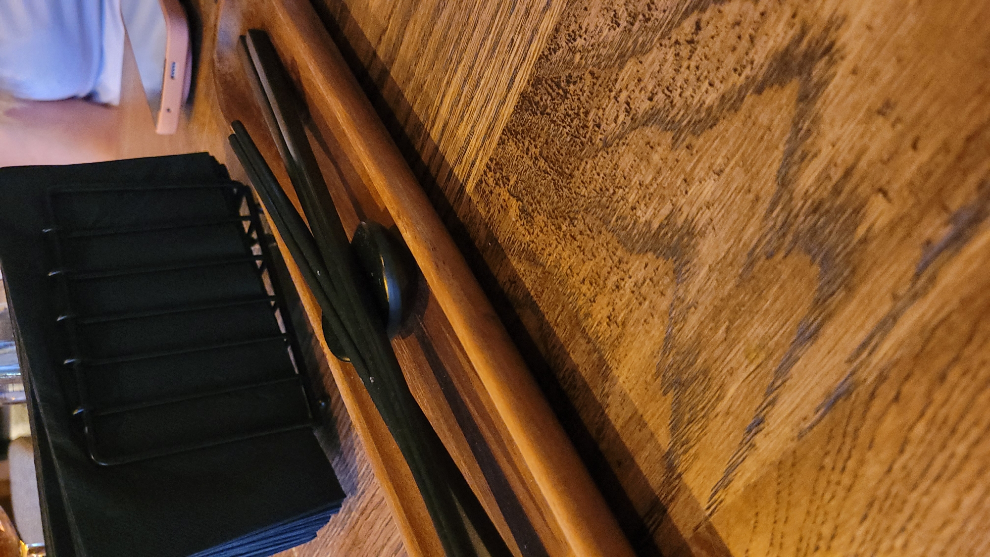 A wooden tray with black chopsticks and a small dark bowl rests on a wooden table. A black napkin holder filled with black napkins is visible in the background, along with a pink smartphone and a person's shoulder and arm. The image appears to be taken in Sofia, Bulgaria.