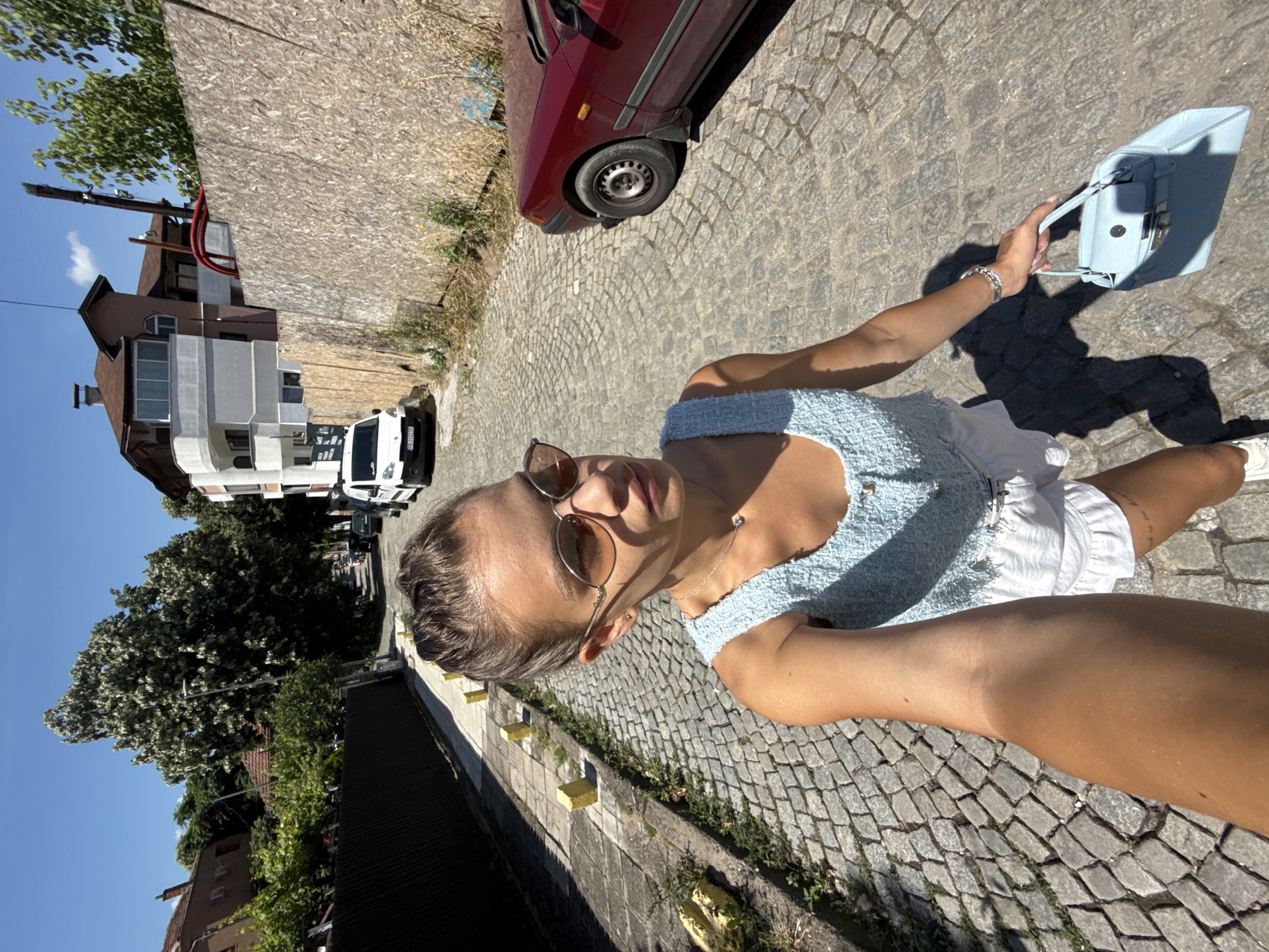 A young woman with her hair pulled back and wearing sunglasses is taking a selfie while walking down a cobblestone street in Plovdiv, Bulgaria. She is wearing a light blue textured tank top and white shorts. She is holding a light blue handbag in her right hand. A red car is parked on the right side of the street, and a white van is visible further down the street. The buildings on either side of the street appear to be residential, with one building under construction or renovation, indicated by the wooden sheeting. The sky is clear and blue, suggesting a sunny day. The street is lined with yellow bollards on the left side.