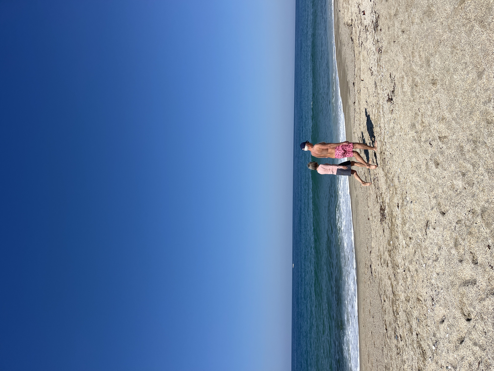 The image shows a beach scene, likely taken in daylight. The sky is clear and blue. The beach is sandy, with visible waves. Two people are walking along the shoreline. The person closer to the viewer is wearing pink shorts and a blue cap. The second person is wearing a pink top and a darker skirt. The image does not contain enough detail to determine if the location is in the city of Alanya, Turkey.