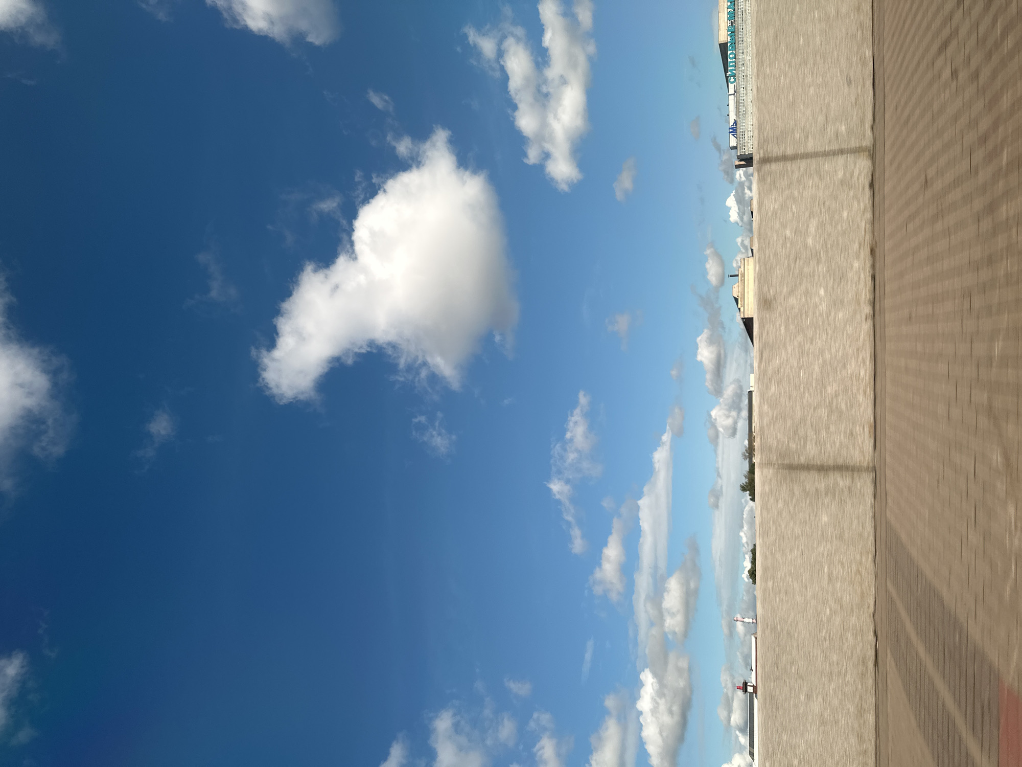 This is a low-angle, wide shot of a bright, sunny day in Saint Petersburg, Russia. The scene is dominated by a vast expanse of blue sky with scattered white clouds. In the foreground, there is a textured, beige concrete wall that extends across the entire width of the image. Below the wall, a patterned brick walkway with shadows suggests movement, possibly from a vehicle. The background reveals a cityscape with several buildings and industrial structures. One building is white with blue lettering that appears to be a Russian company name. Another building has a red and white tower, possibly a smokestack or industrial chimney. The overall impression is one of a bright, daytime scene in an urban or industrial area.