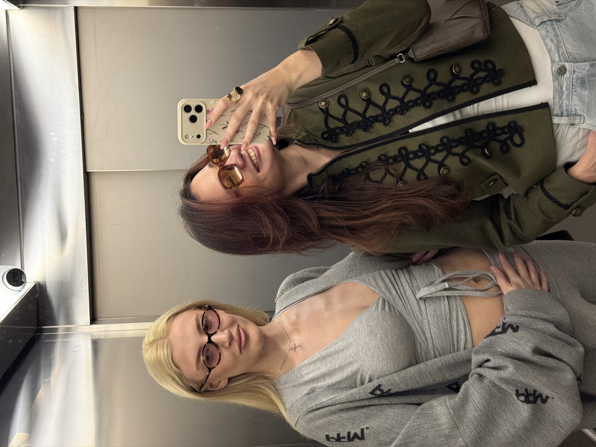 Two young women pose for a selfie in what appears to be an elevator. The woman taking the photo has brown hair, sunglasses, and is wearing a green jacket with decorative black trim. The other woman has blonde hair, glasses, and is wearing a grey outfit. The setting is a small, enclosed space with metal walls, likely the interior of an elevator. The photograph doesn't provide any data to indicate Belgrade, Serbia.