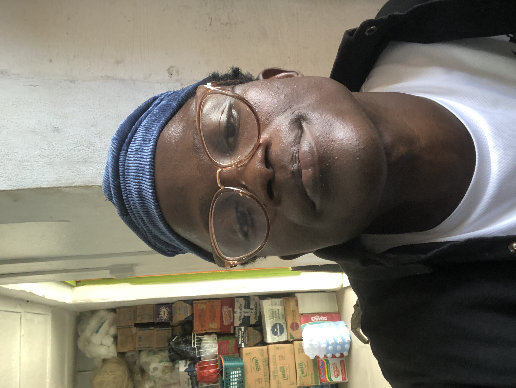 A dark-skinned man wearing a blue beanie and rose-gold framed glasses is shown in a close-up selfie. He is wearing a black jacket over a white t-shirt. The background consists of shelves filled with various boxed products, including what appear to be food and beverage items. To the left, there is a doorway leading to a brightly lit area, possibly an outdoor space. The scene suggests a retail or storage environment, potentially a shop or warehouse, located in Ado, Nigeria.