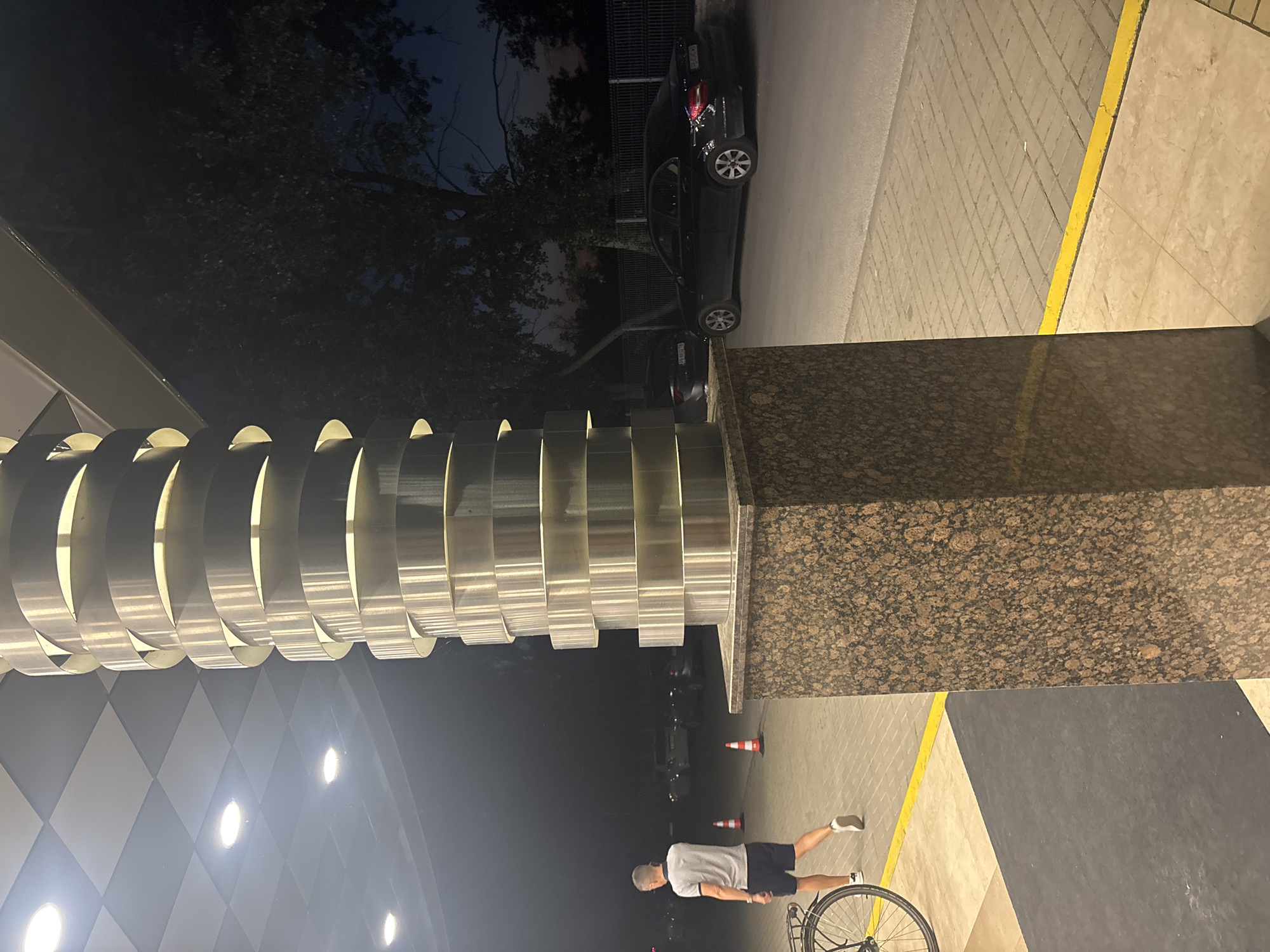 The image shows an outdoor scene at night in Plovdiv, Bulgaria. A tall, decorative, cylindrical sculpture with a series of illuminated, horizontal metal rings dominates the left side. A square column made of granite is next to the sculpture. A man is seen standing next to a bicycle on a sidewalk. The sidewalk also includes orange traffic cones. Cars are parked along the street in the background. The street appears to be paved with light-colored bricks. The sky is dark, with trees visible in the background.