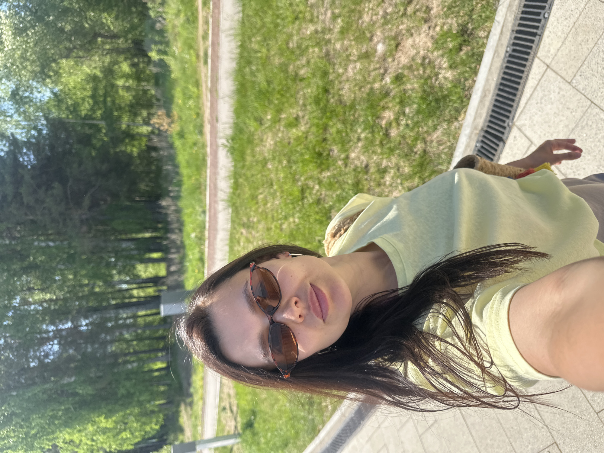 A selfie photograph shows a woman with dark hair and sunglasses wearing a yellow hooded sweatshirt. She is outdoors, sitting near a grassy area with trees in the background. A concrete sidewalk with a drainage grate is visible. The location is in Chaikovsky, Russia.