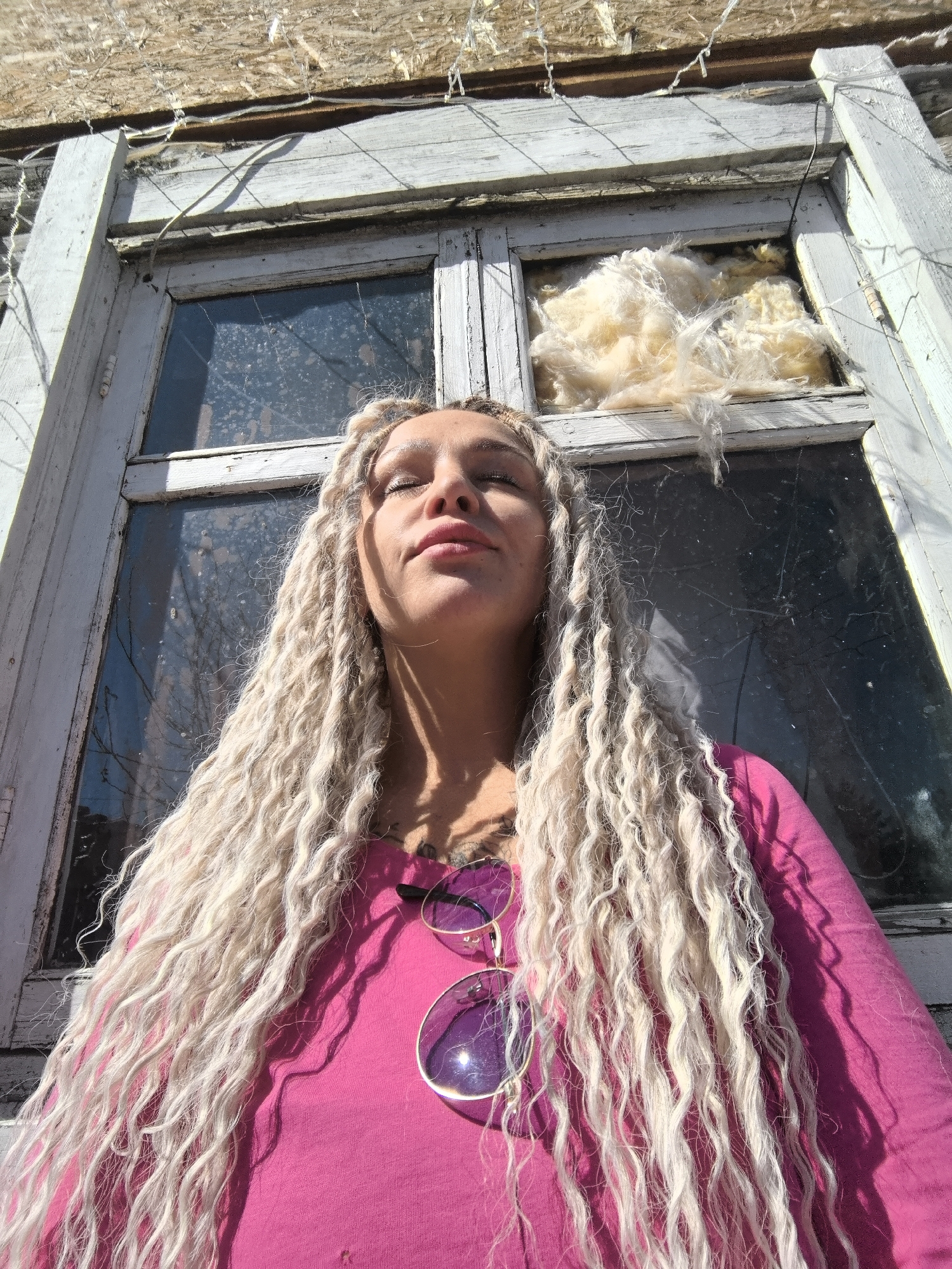 A woman with long, blonde, crimped hair stands against a backdrop of an old, white-framed window, likely in an urban setting based on the architecture. The woman is wearing a pink long-sleeved shirt and has a pair of sunglasses hanging on her shirt. The window appears to be partially broken, with exposed insulation visible. The setting might be Noviy, Russia, based on user instructions, but specific geographical details are not apparent in the image itself. The woman's eyes are closed, and she is facing the sun.