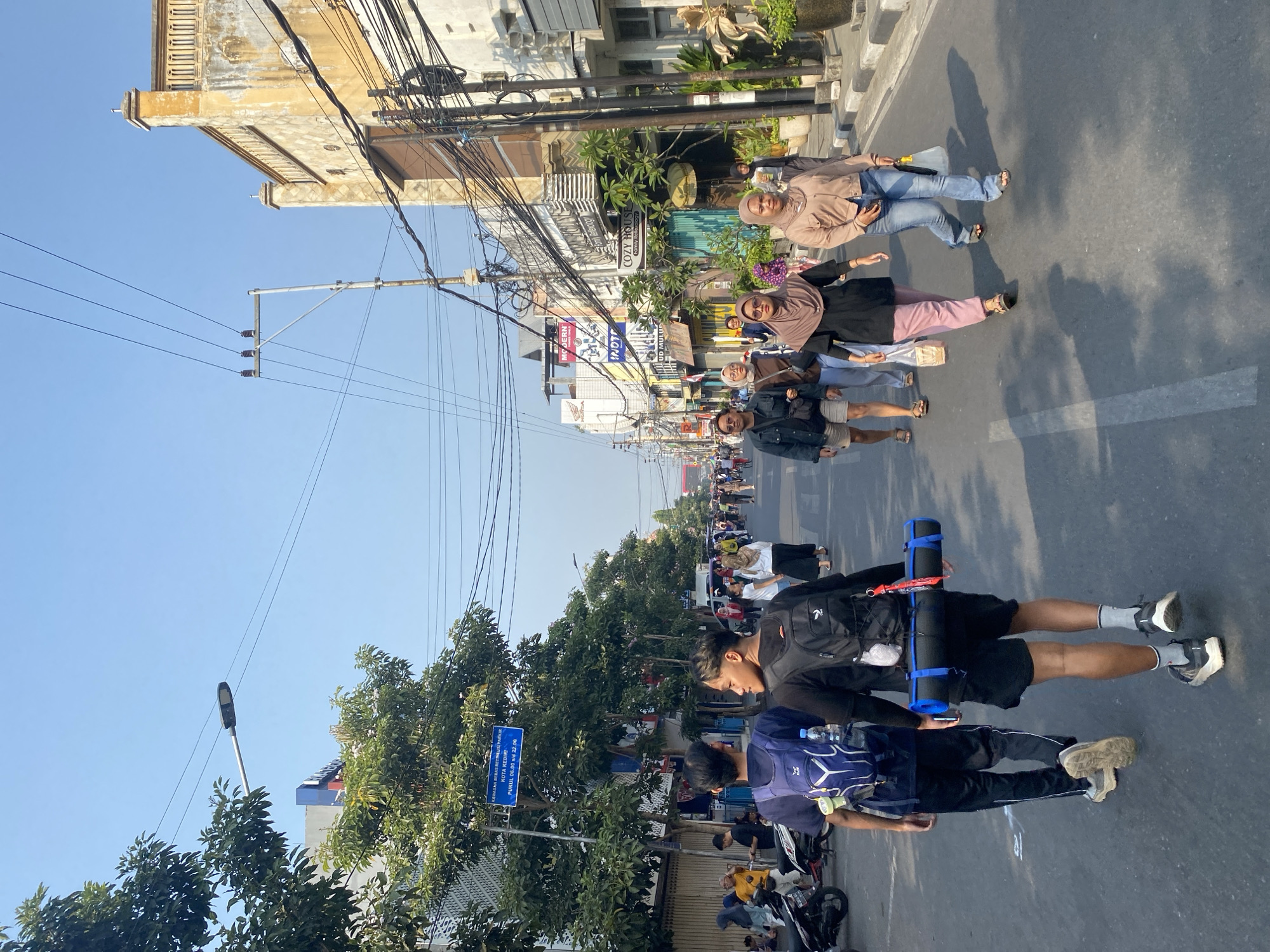The image captures a street scene in the city of Kediri, Indonesia, characterized by buildings, a road, a sidewalk, and several individuals. The sky is clear and blue, indicating fair weather. On the left side of the street, lush green trees line the sidewalk, their foliage casting shadows on the road. Buildings of varying architectural styles are visible along the street, some appearing older with ornate decorations and faded paint, while others are more modern. Overhead, a complex network of electrical wires and utility poles crisscrosses the sky, a common sight in urban environments.
Several people are walking on the street and sidewalk. In the foreground, two young men are walking away from the camera. The man on the left wears a dark blue backpack and black pants. The man on the right, also with a backpack, carries a rolled-up sleeping mat horizontally across his back. Further down the street, a group of people, including women wearing headscarves and jeans, are walking together. One woman is dressed in a black top and pink pants, while another wears a beige jacket and blue jeans. Other individuals are seen walking in the distance, as well as parked motorcycles and scooters on the left side of the street. The road surface is asphalt, marked with white lines. The overall atmosphere suggests a bustling day in a commercial or residential area of Kediri.