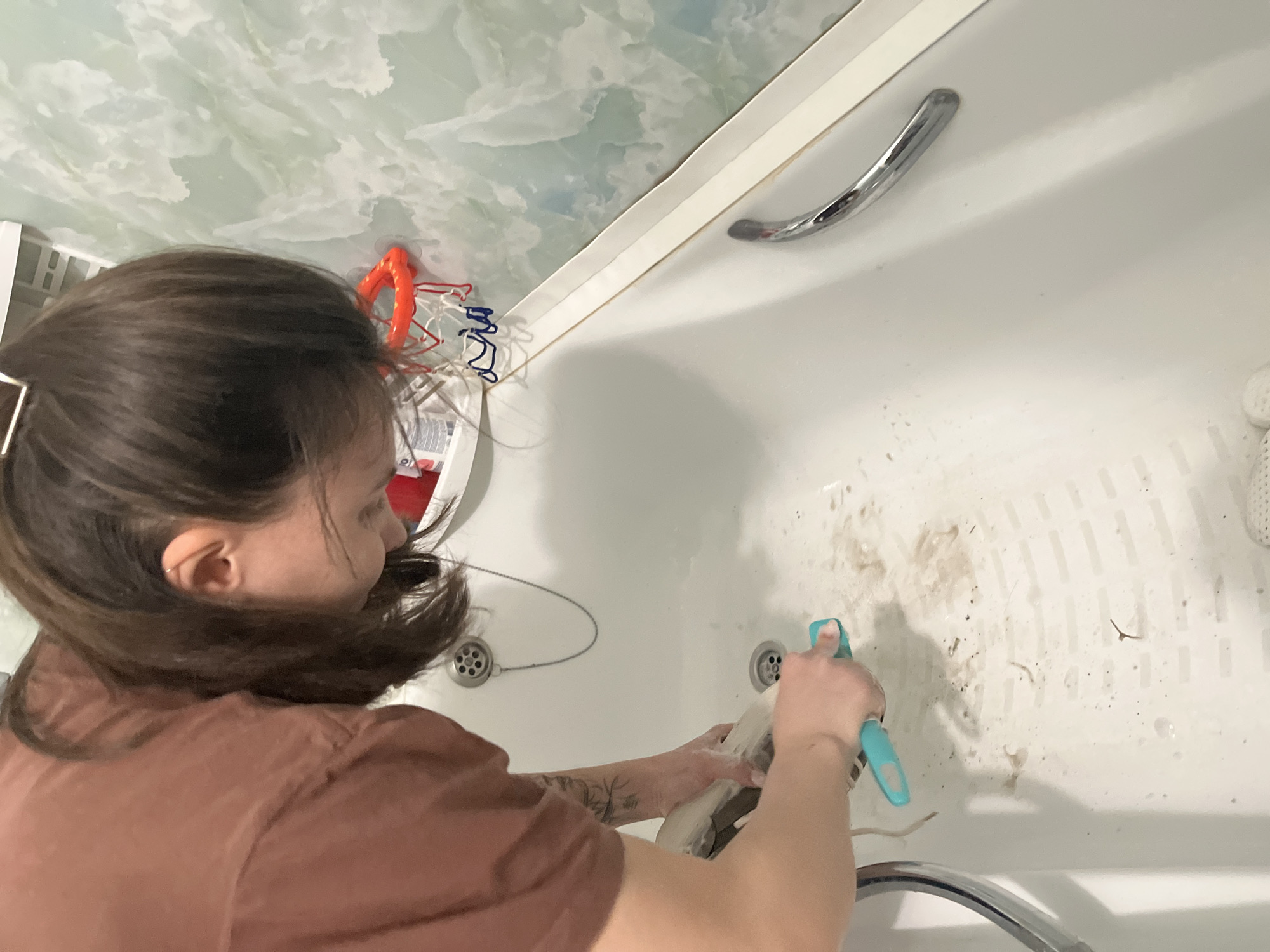 A woman is cleaning a white bathtub. The bathtub has a silver grab bar and a chrome faucet. There is debris in the bathtub, including hair and dirt. The woman is holding a spray bottle in her right hand and is spraying the bathtub. The woman is wearing a brown shirt. The background is a patterned wall. There is an orange basketball hoop hanging near the edge of the bathtub.
I have no information about the location being in the city of Oryol-Izumrud, Russia, so I have omitted it from the description.