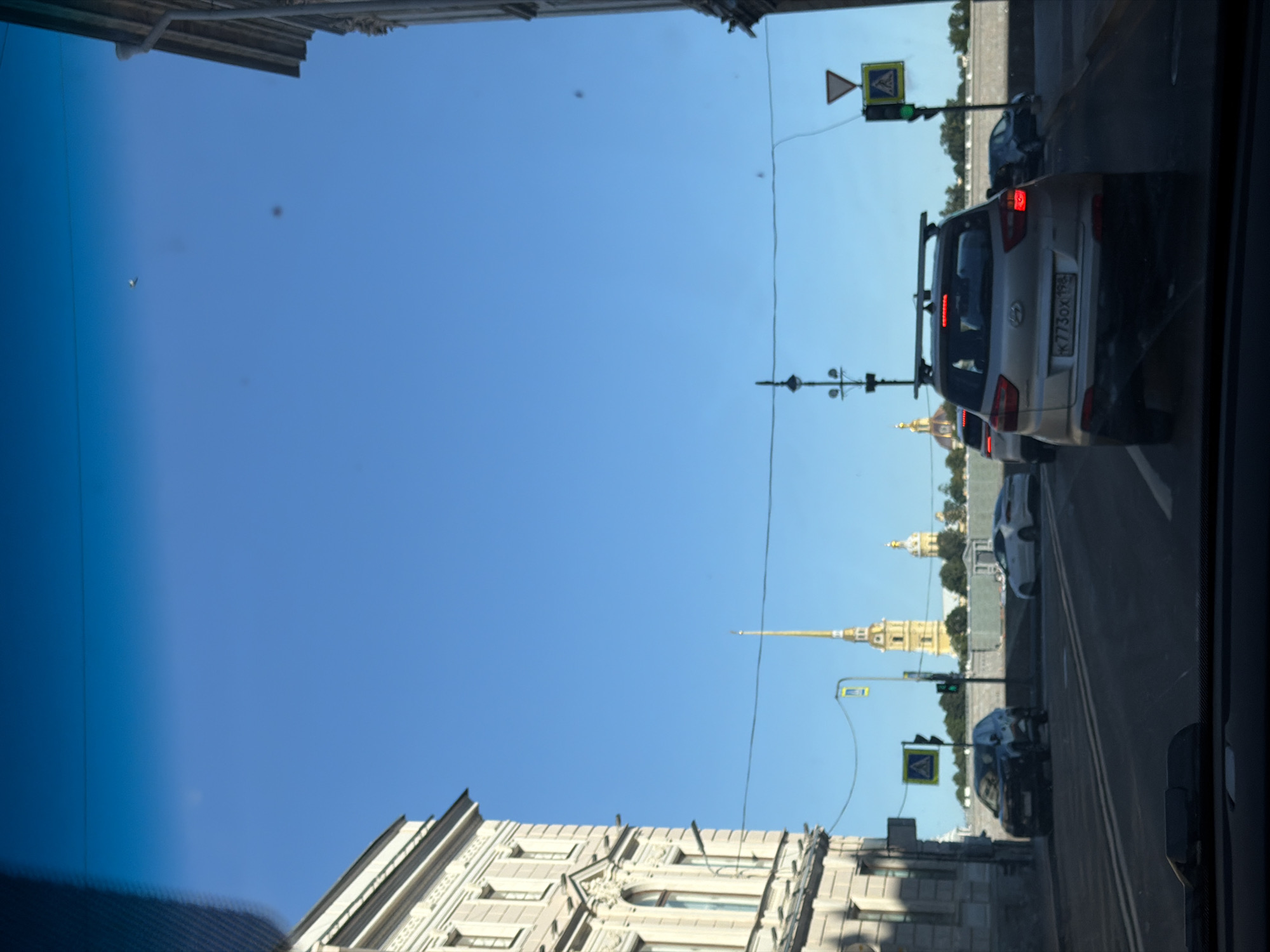 The image shows a view from inside a car, looking out at a city street in Saint Petersburg, Russia. The sky is clear and blue. On the left is a light-colored building with ornate details. In the distance, a tall, golden spire of a church or cathedral is visible, along with other church domes. Several cars are on the road, including a silver SUV in the foreground with a license plate that reads "K773OX 198". Traffic lights and street signs are also present. The overall scene depicts a sunny day in an urban environment.