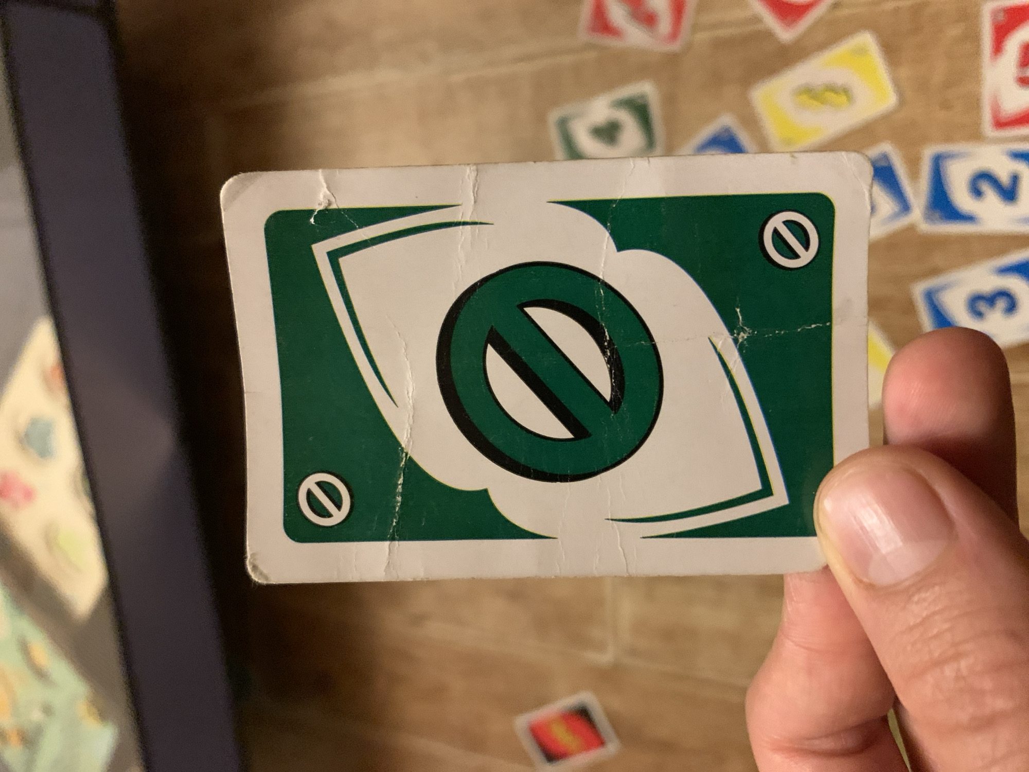 A close-up, slightly overhead shot shows a person's hand holding a green Uno "Skip" card. The card is positioned vertically in the center of the frame, with the person's thumb and index finger obscuring the bottom left corner. The card has a white border and features a large, stylized white shape with a black outline in the center, enclosing a black circle with a diagonal line through it. Two smaller black circles with diagonal lines through them are located in the top left and bottom right corners of the card. The card appears to be slightly creased and worn, with small tears along the edges. The background consists of a wooden surface with other Uno cards scattered across it, some visible in focus and others blurred. Among the visible cards are a red card with a yellow symbol, a green card with a white symbol, a yellow card with a green number 3, and blue cards with white numbers 3 and 2. The lighting is warm and direct, casting subtle shadows. The overall scene suggests a casual game of Uno in progress.