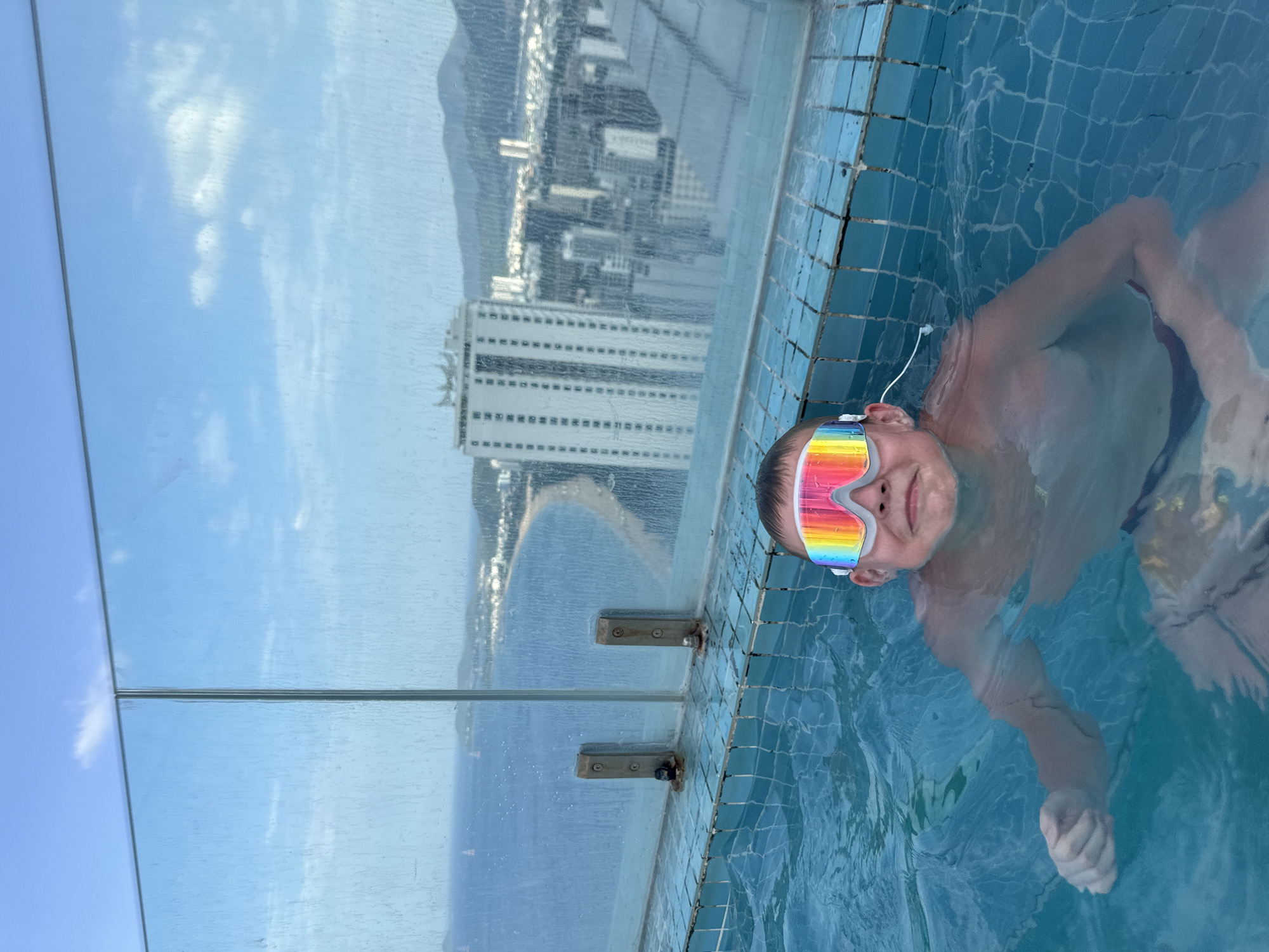 A young boy with rainbow-striped swimming goggles and a white earbud is seen smiling and floating in a swimming pool. His body is partially submerged in the clear blue water, which has blue mosaic tiles along the edge. The pool appears to be a high-rise or rooftop pool, featuring a transparent glass barrier that offers a panoramic view.
Beyond the pool and glass barrier, a sprawling cityscape unfolds, with numerous tall buildings stretching towards a curved coastline. A sandy beach separates the city from the vast blue ocean. Mountains are visible in the distant background, completing the scenic vista. The sky is bright and clear, with some scattered white clouds reflected in the glass. The scene takes place during the daytime under sunny weather. The overall impression is one of leisure and enjoyment in an impressive high-altitude setting, likely overlooking Nha Trang, Vietnam. No visible text is present in the image.