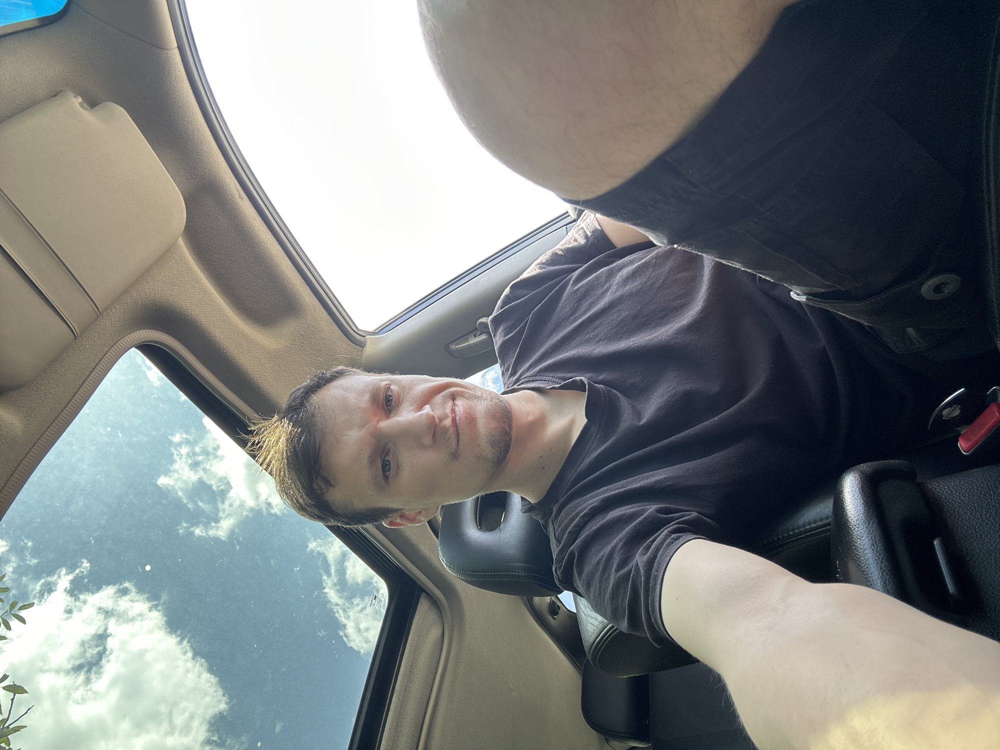 A man is sitting in the driver's seat of a car, looking towards the camera. The car has a sunroof that is open, revealing a cloudy sky. The man is wearing a black t-shirt and dark shorts. He has short, light brown hair and is clean-shaven. The interior of the car appears to be a dark color, possibly black leather. The image was taken outdoors during the daytime. There is no indication that this image was taken in Makiv, Ukraine.