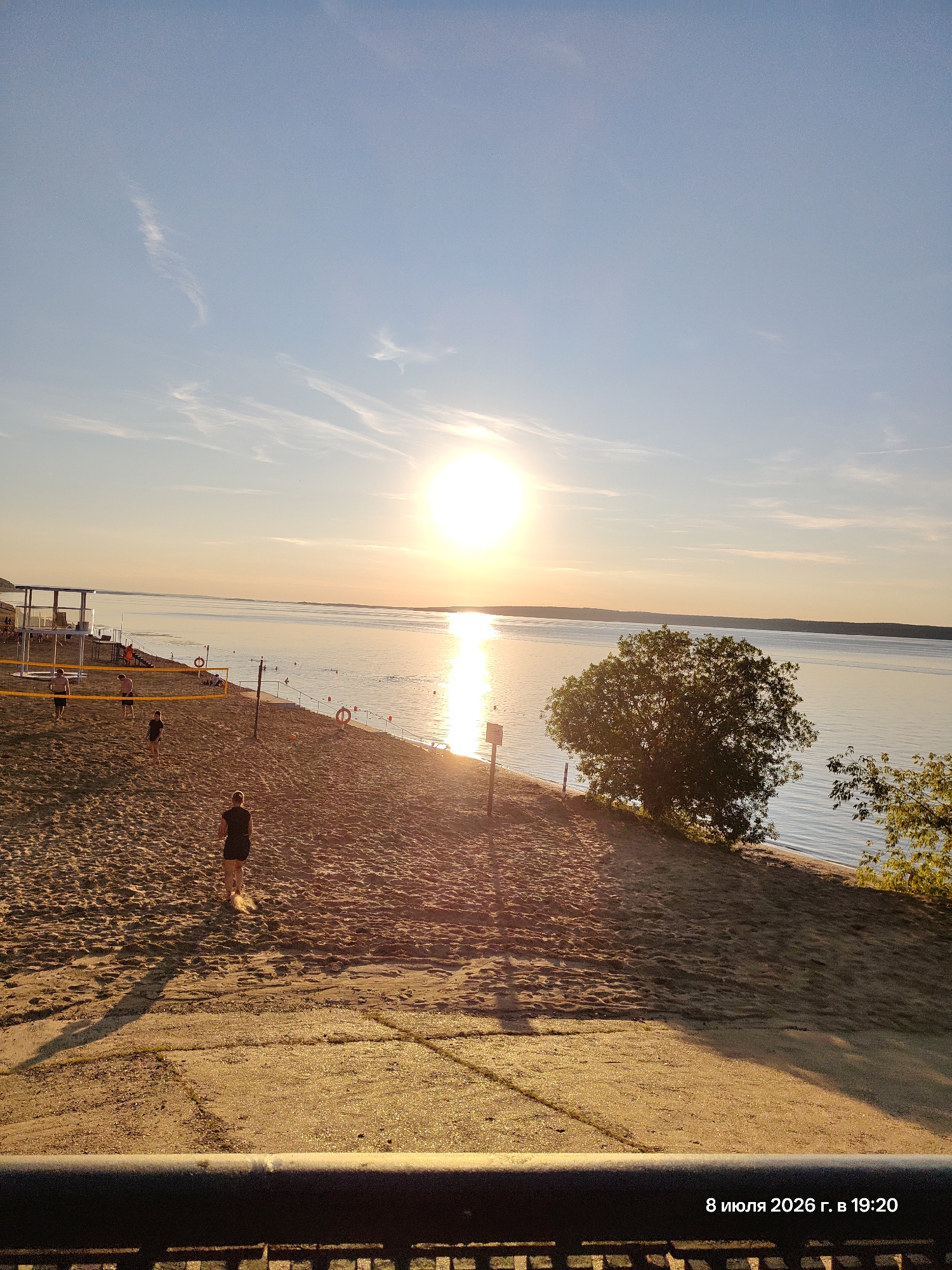 This is a landscape photograph of a beach at sunset. The sun is low in the sky, casting a warm glow over the scene. The sky is a clear blue with some wispy clouds. The beach is made of sand and there are several people playing volleyball. The water is calm and reflects the colors of the sunset. There is a tree on the right side of the image, and some foliage in the foreground. The date and time stamp at the bottom right corner reads "8 июля 2026 г. в 19:20".