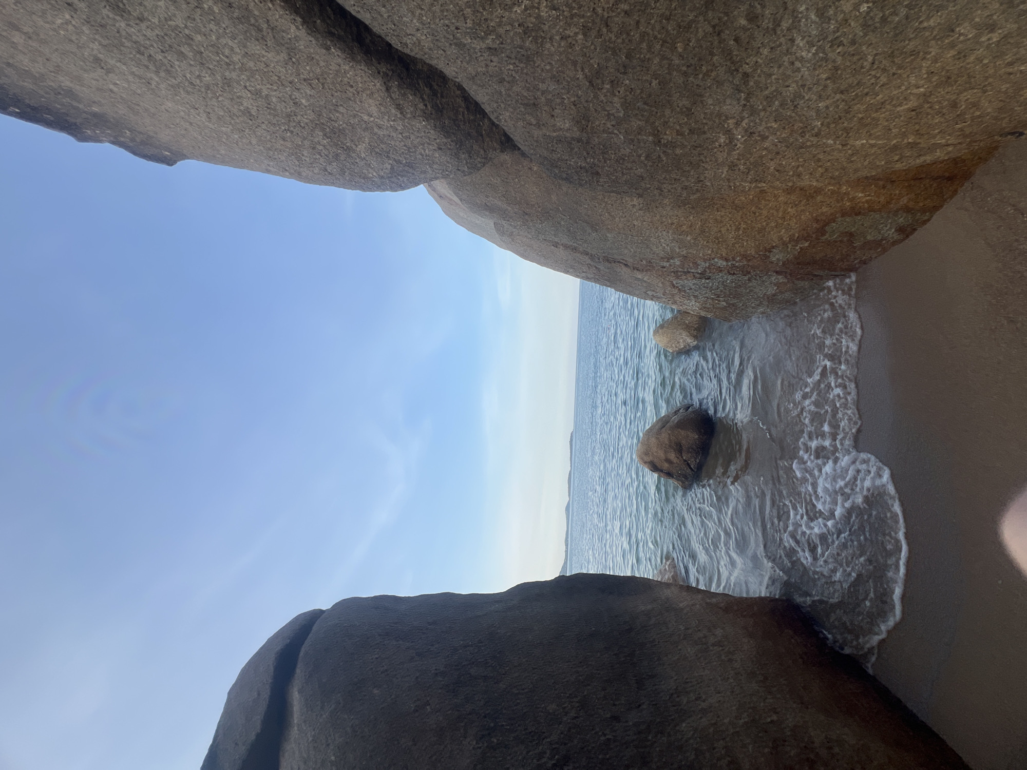 A low-angle, wide shot captures a serene beach scene in Ko Samui District, Thailand. Two large, weathered granite boulders frame the foreground, their rough surfaces textured with shades of grey and brown. Between these natural pillars, the gentle, blue ocean stretches towards the horizon, where a faint, hazy landmass is visible. Small waves lap at a sandy shore, creating frothy white foam. A few medium-sized, rounded rocks emerge from the shallow water. The sky above is a clear, pale blue, with subtle wisps of clouds. The overall impression is one of tranquility and natural beauty.