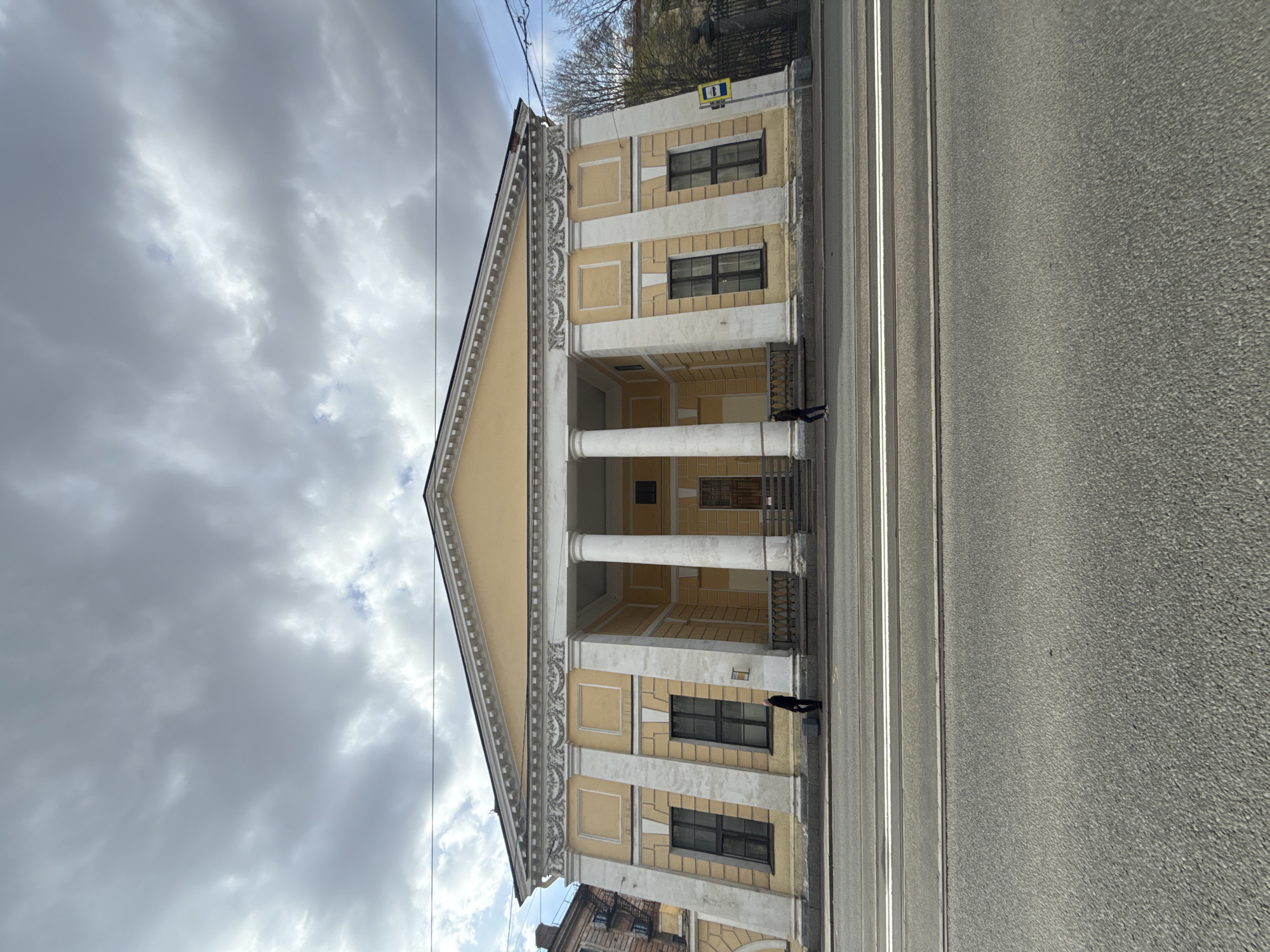 A grand, classical-style building with a light yellow facade and white architectural details stands on the left side of a wide city street. The building features a prominent portico with four large, white fluted columns supporting a triangular pediment. Its facade is adorned with decorative elements and multiple rectangular windows with dark frames.
On the right, a broad asphalt road occupies the foreground, traversed by parallel tram tracks marked with white lines. Two individuals are visible on the sidewalk near the building; one near the left edge of the frame and another closer to the portico columns, appearing to walk or stand.
The sky above is heavily overcast with dramatic grey clouds, indicating a cloudy daytime. Bare tree branches are visible behind the building on the far right, suggesting a colder season. A small rectangular yellow sign with blue elements, possibly a white symbol on a blue background, is affixed to the upper right corner of the building's facade, though its text is not legible.
The scene captures a typical urban environment, with the imposing architecture and tram tracks being characteristic of Saint Petersburg, Russia.