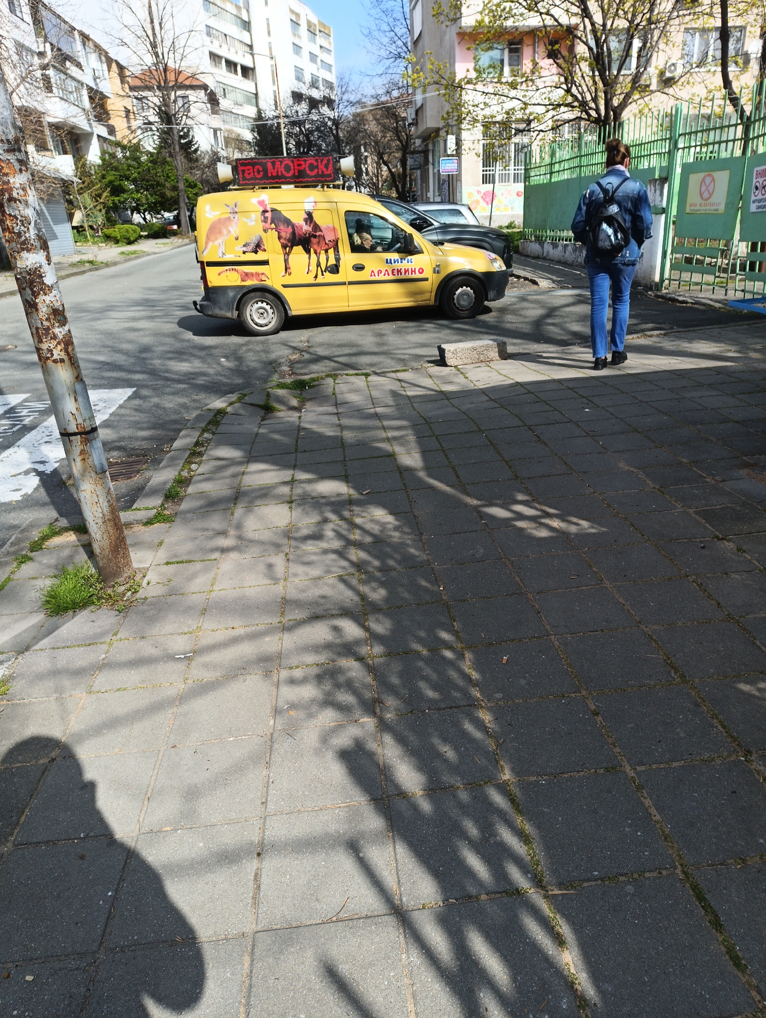 The image shows an outdoor scene in what appears to be a residential area of Burgas, Bulgaria. A yellow van with animal circus graphics and the text "Circus Draklino" and "Circus Morski" printed on it is parked on the left side of the road. The van is parked behind a person walking on a paved sidewalk. The person is wearing jeans and a jean jacket and is carrying a backpack. A rusty utility pole is located next to a crosswalk and sidewalk in the foreground. Behind the van, other parked cars are visible, as well as buildings, trees, and a fence. The sidewalk is paved with square concrete blocks. The time of day appears to be mid-day, evidenced by the shadows cast on the sidewalk. The location is likely near a school or public space due to the presence of the fence.