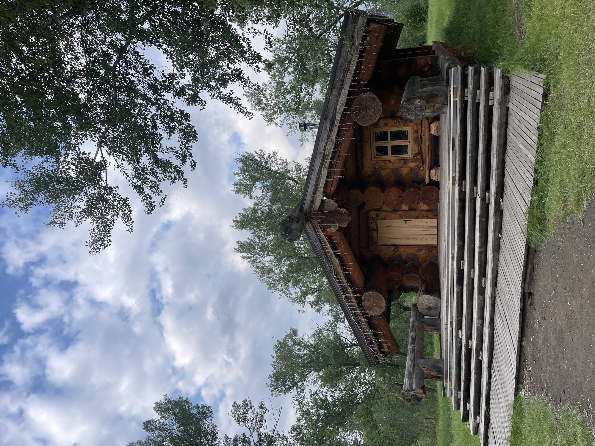 A rustic, wooden log cabin with a large, multi-tiered wooden deck and steps sits nestled amongst lush green trees under a cloudy sky. The cabin has a prominent wooden door and a small window with a grid pattern. Decorative string lights are draped along the eaves of the cabin's roof. Two large log benches are situated on the deck, one on either side of the entrance. The area surrounding the cabin is covered in grass, with a dirt path leading up to the deck. This scene is located in the city of Krasnoyarsk, Russia.