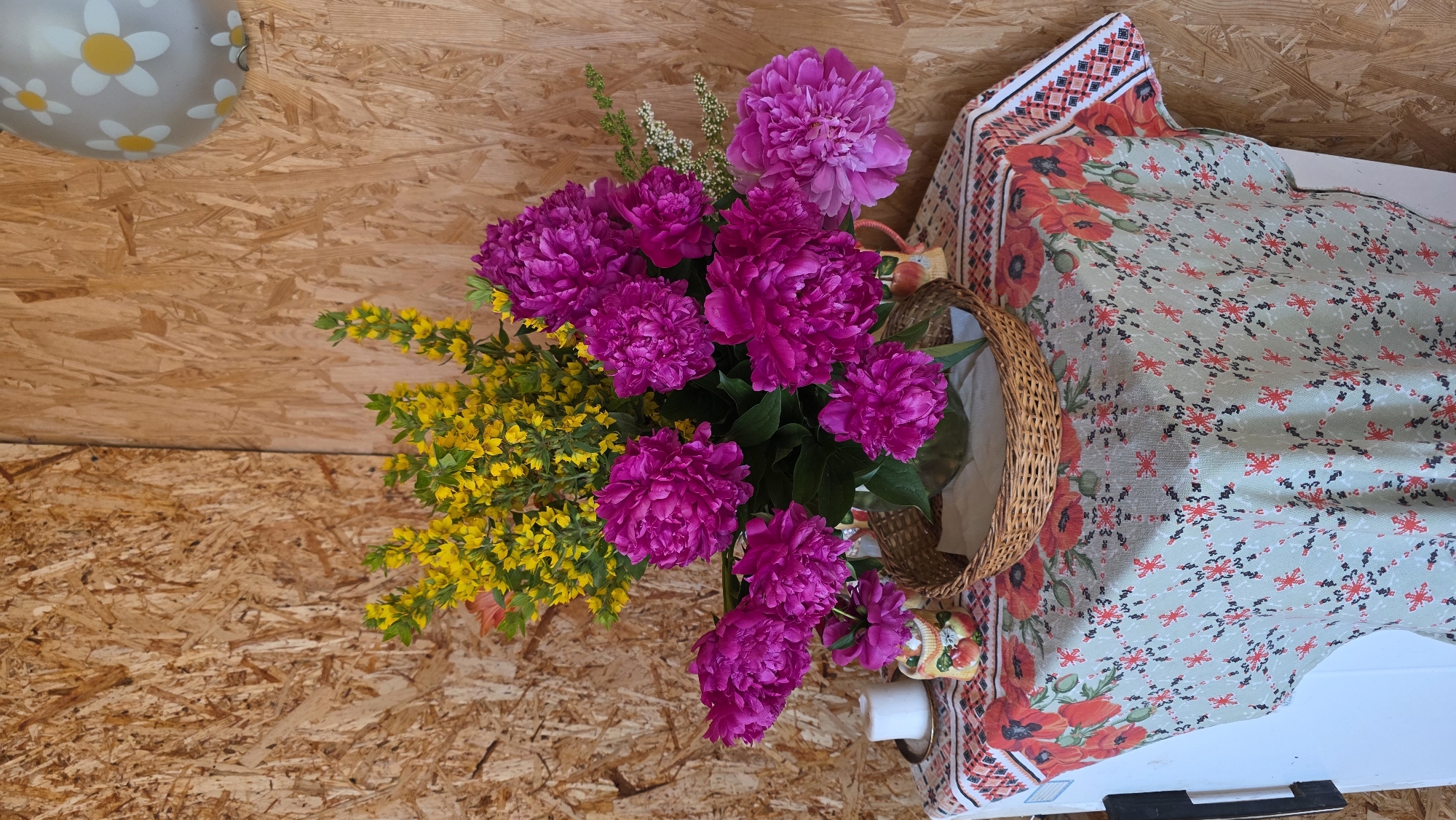 A vibrant still life features a bouquet of fuchsia peonies and yellow flowers, arranged in a glass vase within a woven basket. The bouquet rests on a table covered with a tablecloth adorned with a red floral and patterned design. To the left, a white candle sits on a small saucer next to a decorative ceramic jug depicting apples. In the background, a rustic wooden wall provides a textured backdrop, with a portion of a gray balloon featuring white and yellow daisy patterns visible in the upper right corner. The scene is likely set indoors, possibly in a rural home or a studio space in the village of Kupoyarovo, Russia.