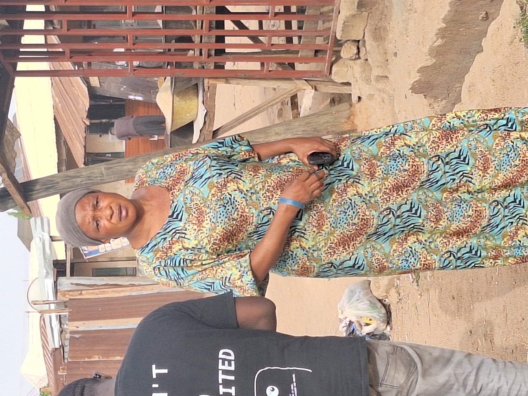 In Sabon Tasha, Nigeria, an older woman in a patterned blue, brown, and yellow dress and a grey head covering is holding a small black electronic device. She appears to be looking at the camera. A portion of a man wearing a black t-shirt and light-colored pants is visible in the foreground. The background includes corrugated metal structures and a stone wall. A red metal framework is overhead. A plastic bag of debris is nearby.