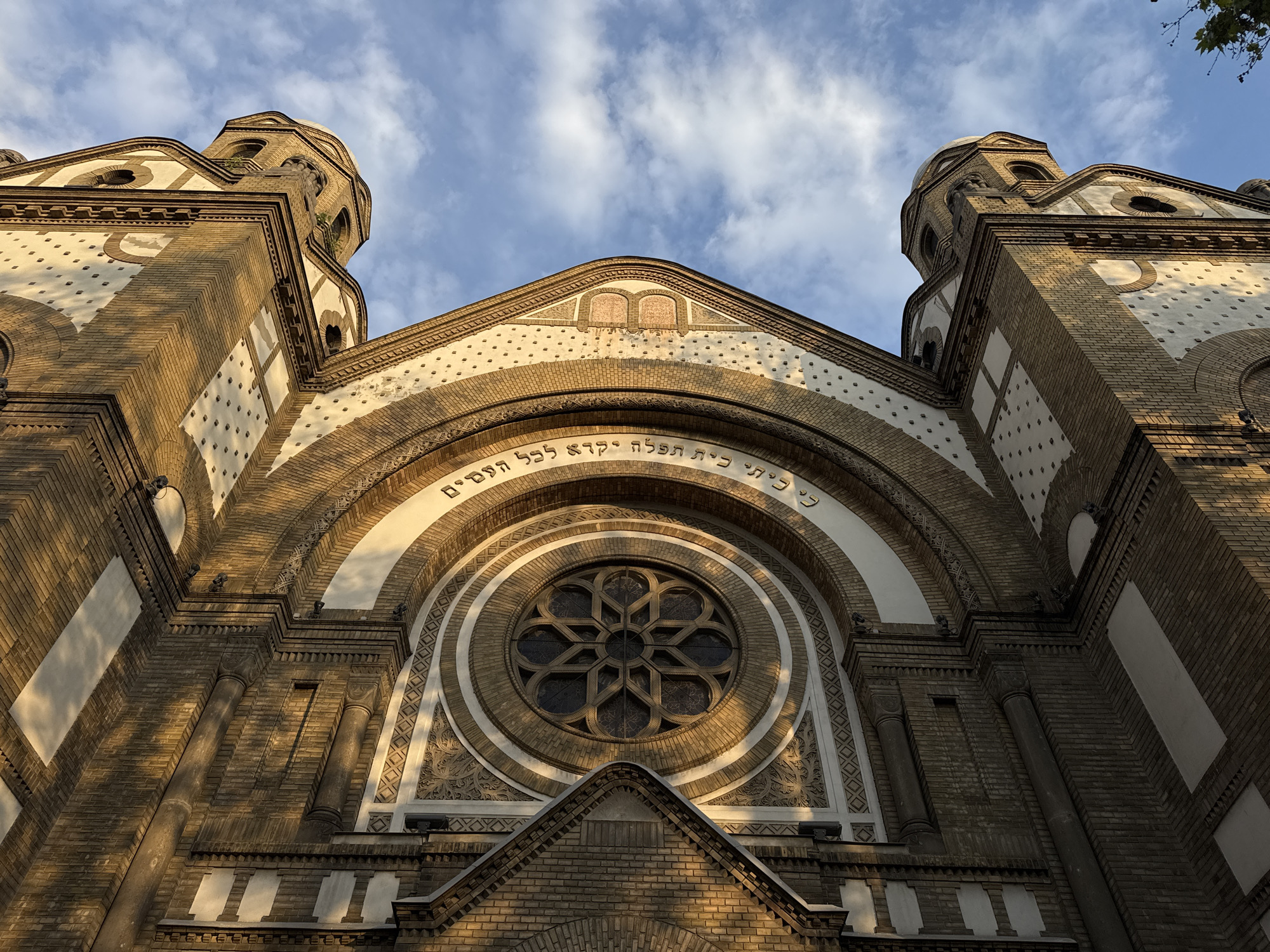 A low-angle shot captures the facade of the Novi Sad Synagogue in Novi Sad, Serbia. The building, made of yellow brick with white and cream accents, features two towers on either side of a central structure. A large rose window dominates the facade. An inscription in Hebrew script arcs over the rose window. The sky above is blue with scattered white clouds.