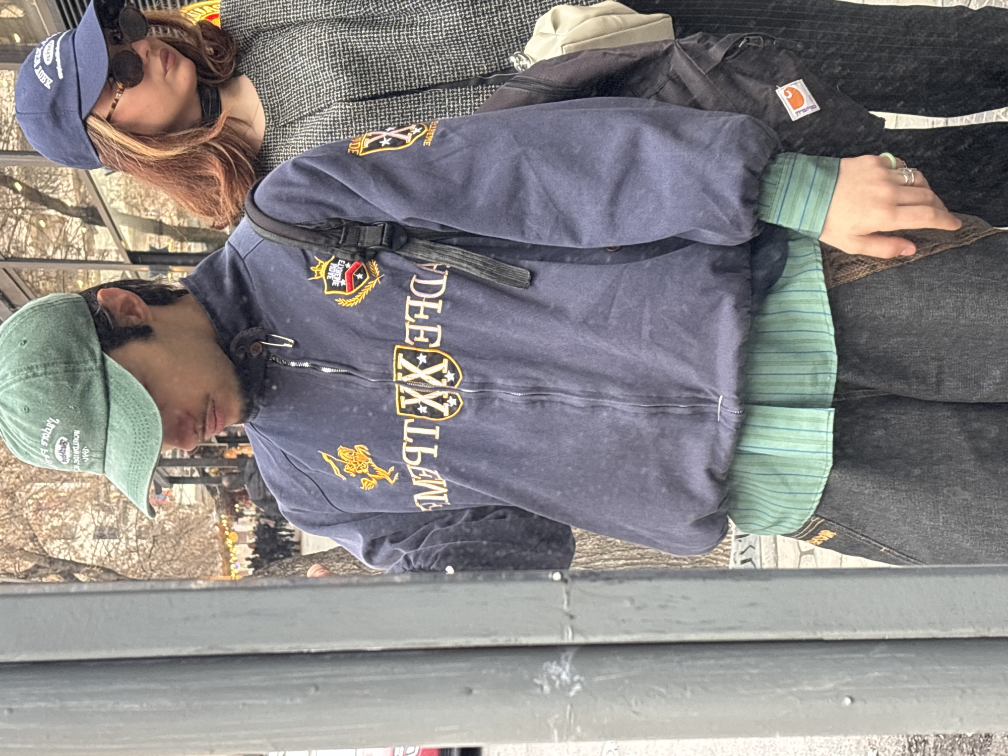 Two individuals are visible, reflected in a window. One wears a navy blue baseball cap, sunglasses, and a grey patterned jacket. The second person is wearing a green baseball cap and a blue zip-up jacket with decorative patches and text, and a green striped shirt underneath. They are in an urban setting, likely Yerevan, Armenia, as suggested by the presence of street lights and possible architectural styles reflected in the glass.