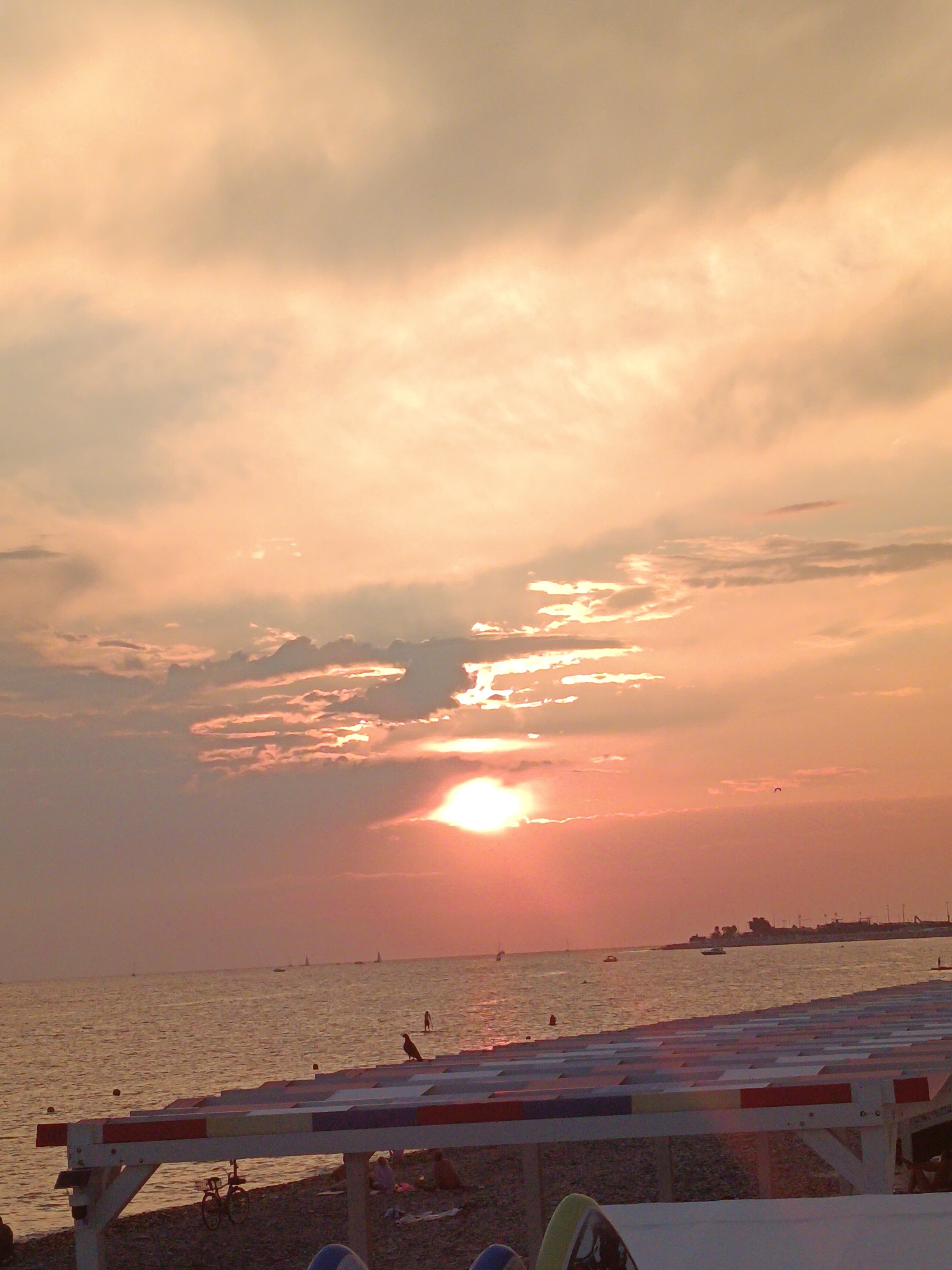 The image shows a beach scene at sunset in the city of Sirius, Russia. The sun is low in the sky, casting a warm orange glow over the clouds and the water. The sea is calm, with small waves lapping at the shore. In the foreground, there is a white structure with colorful stripes, possibly part of a beach umbrella or a seating area. A bicycle is parked underneath this structure, and a bird is perched on top of it. Further out in the sea, several sailboats and a distant coastline can be seen. There are also a few figures on the beach and in the water, suggesting the presence of people enjoying the sunset.
