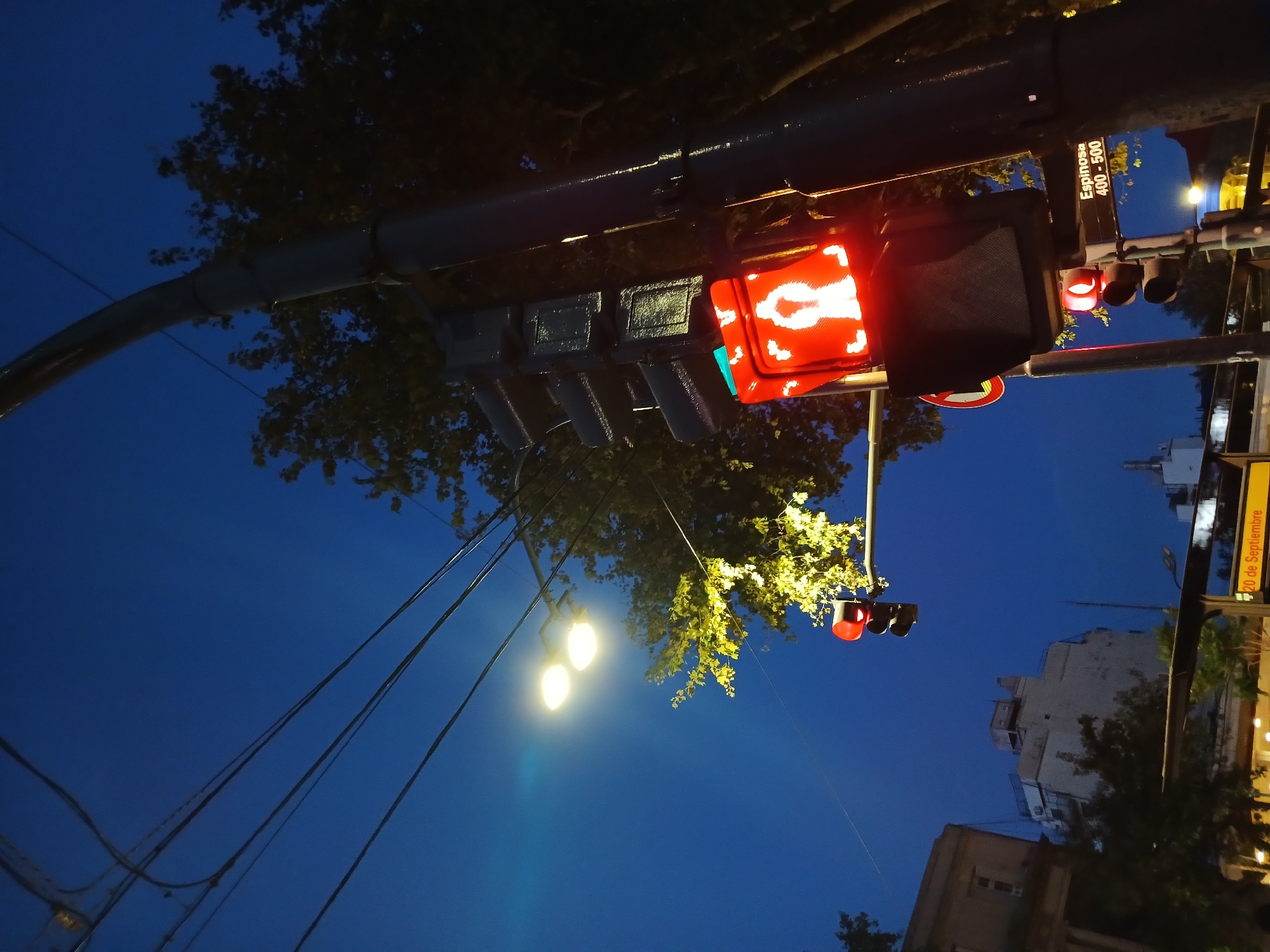 The image depicts a low-angle view of a street scene in Buenos Aires, Argentina, at night or dusk. The dominant element is a traffic light signal with a red pedestrian crossing symbol illuminated. Adjacent to the pedestrian signal is a standard traffic light also showing a red light. Above the traffic lights is a street sign that reads "Espinosa 400-500". Power lines and bare tree branches are visible against a deep blue sky. A street light shines brightly in the distance. In the background, buildings can be partially seen, along with another sign indicating "20 de Septiembre". The overall scene suggests a quiet urban setting at twilight or in the early evening.