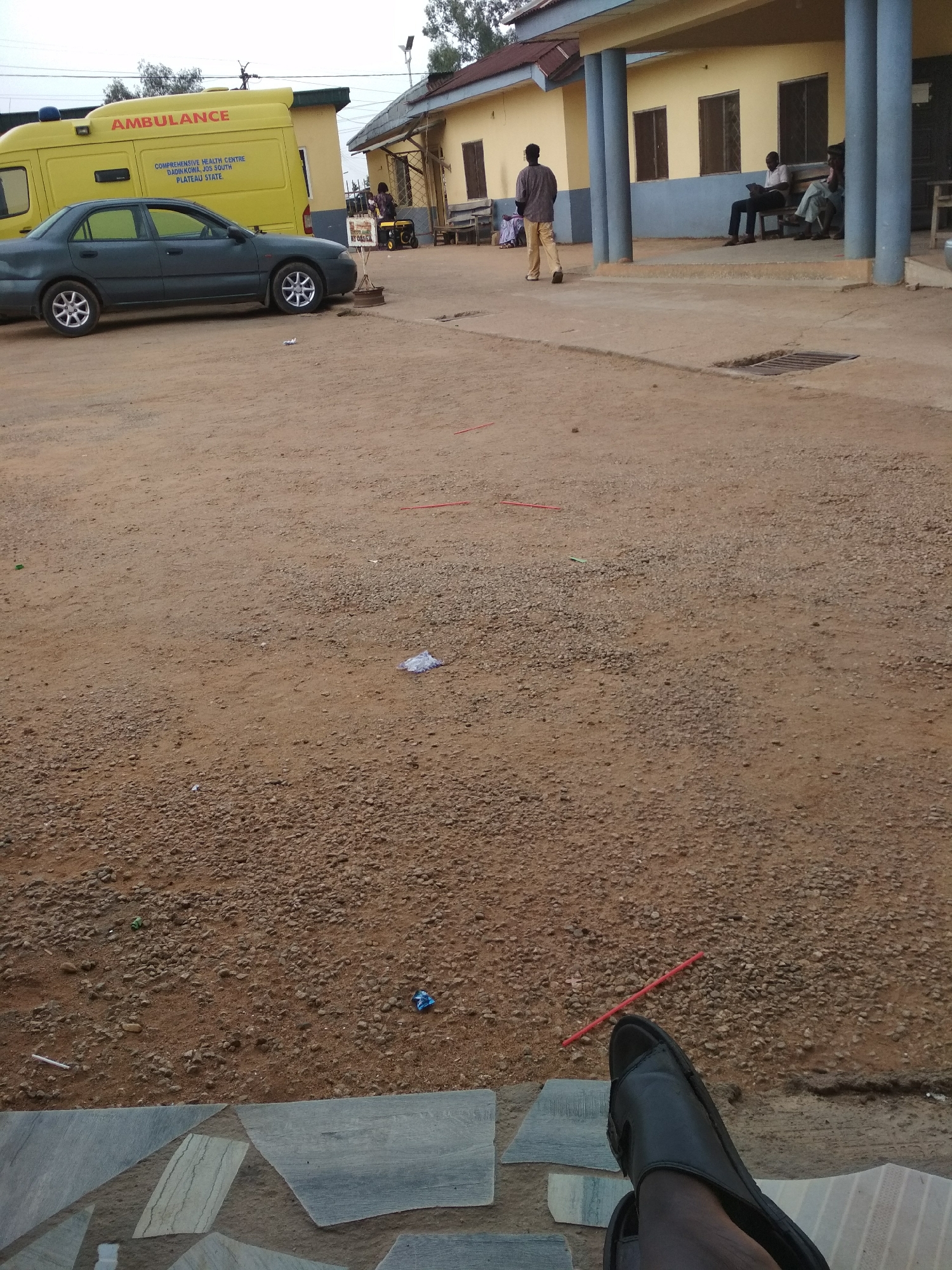 The image depicts an outdoor scene at what appears to be a medical facility or hospital, possibly in an area with limited resources. A yellow ambulance is parked next to a gray car. A building is in the background with a veranda, where a few people are sitting. A man sweeps the yard. The ground is dirt and gravel. A pair of sandals are in the foreground. There is litter scattered on the ground. The location appears to be in the city of Unknown, Unknown.