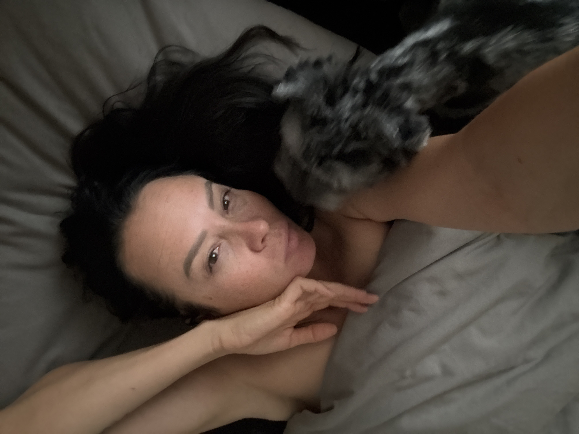 A top-down, close-up shot shows a woman lying in bed with a gray comforter. Her dark hair is spread across the pillow, and one arm is resting on her forehead, while the other arm is visible in the lower left corner, partially obscured by what appears to be a furry object. The woman has brown eyes and fair skin with freckles. Her gaze is directed upwards, and her expression is neutral. The lighting is dim, creating a soft, intimate atmosphere. There is no visible indication of the specific location in the city of Chaikovsky, Russia.