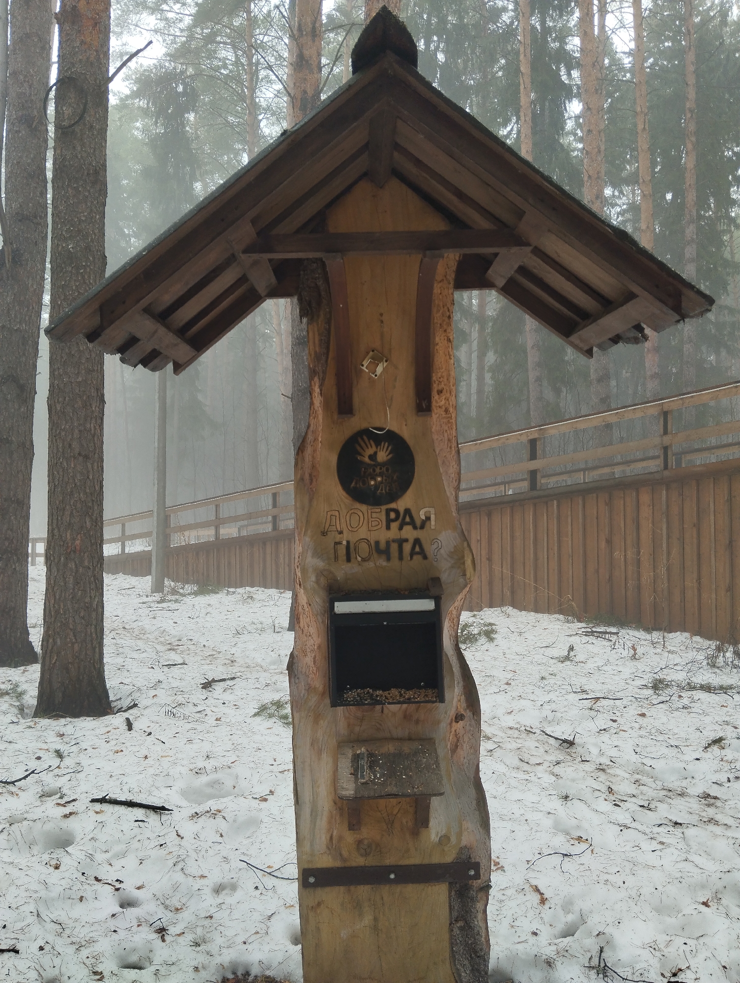 The image depicts a wooden bird feeder shaped like a traditional Slavic-style postbox. It is located in a forest or park setting with snow covering the ground. In the background, pine trees are visible, and there is a wooden fence. The bird feeder has a roof to protect the food from the elements. The feeder itself contains birdseed. An inscription in Russian on the wood reads "Добрая почта?", which translates to "Good mail?" or "Kind mail?". It is likely situated in Votkinsk, Russia.