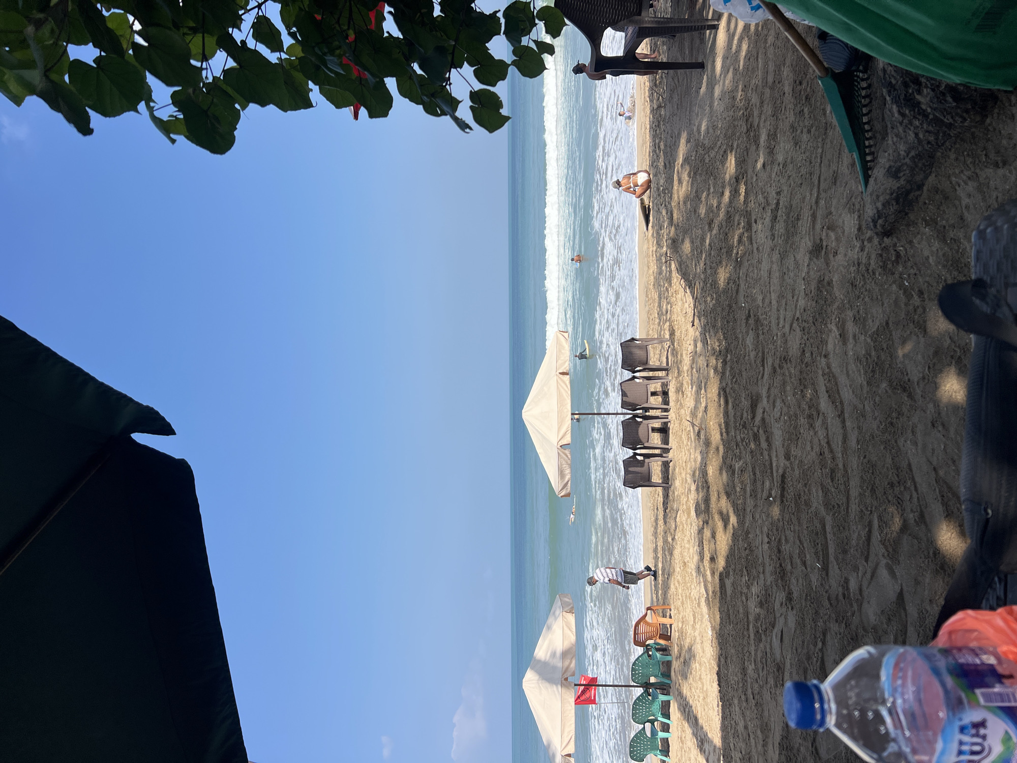 In Kuta, Indonesia, a beach scene unfolds under a clear blue sky with gentle waves lapping the shore. The foreground is dominated by dark sand, with a partially visible plastic water bottle and a green rake hinting at recent activity. Moving towards the water, several beach chairs and umbrellas offer shade. To the left, two beige umbrellas stand over a row of colorful plastic chairs, with a red flag planted in the sand. A man in a striped shirt walks near the water's edge. Further out in the ocean, several individuals are visible, some on surfboards, enjoying the water. To the right, a woman in a white bikini sits on the sand, facing the ocean, with a surfboard beside her. Another person is partially submerged in the water nearby. Dark, woven chairs are also positioned on the sand, and the edge of lush green foliage frames the right side of the image. The overall impression is a peaceful day at the beach, with people engaged in various leisurely activities.