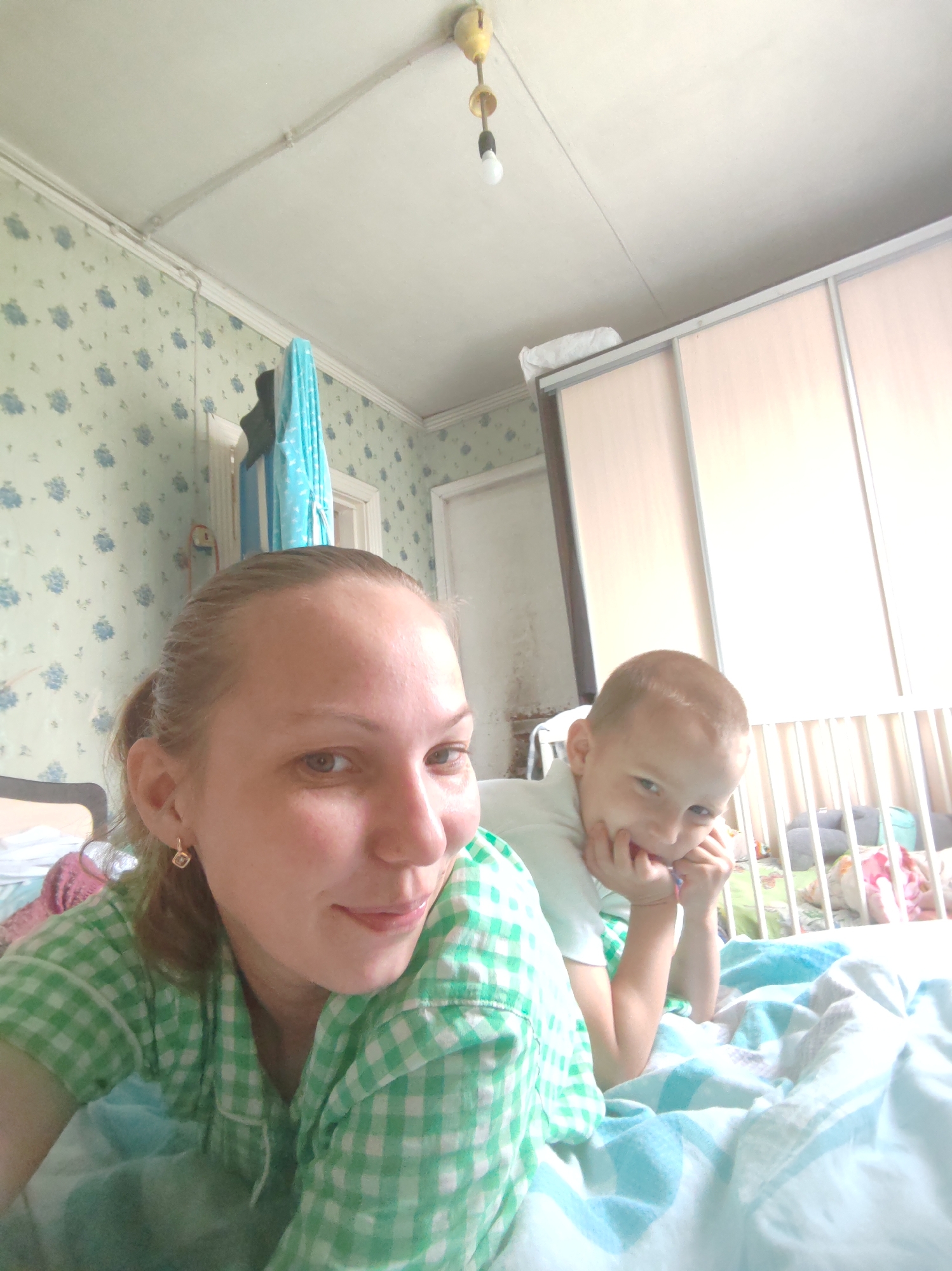 A selfie captures a woman and a young boy lying on a bed in a room located in Чухлэм, Russia. The woman, positioned closer to the camera, is wearing a green and white checkered shirt, revealing a small, square earring on her left ear. She is smiling slightly at the camera. Behind her and slightly to the right, a young boy rests his chin on his hands, also looking at the camera. The bed is covered with a blue and white patterned blanket. In the background, there is a white crib with various objects and toys inside. Behind the crib, a large closet with sliding doors is visible. The walls of the room are covered in a blue floral wallpaper, and a light fixture hangs from the ceiling.