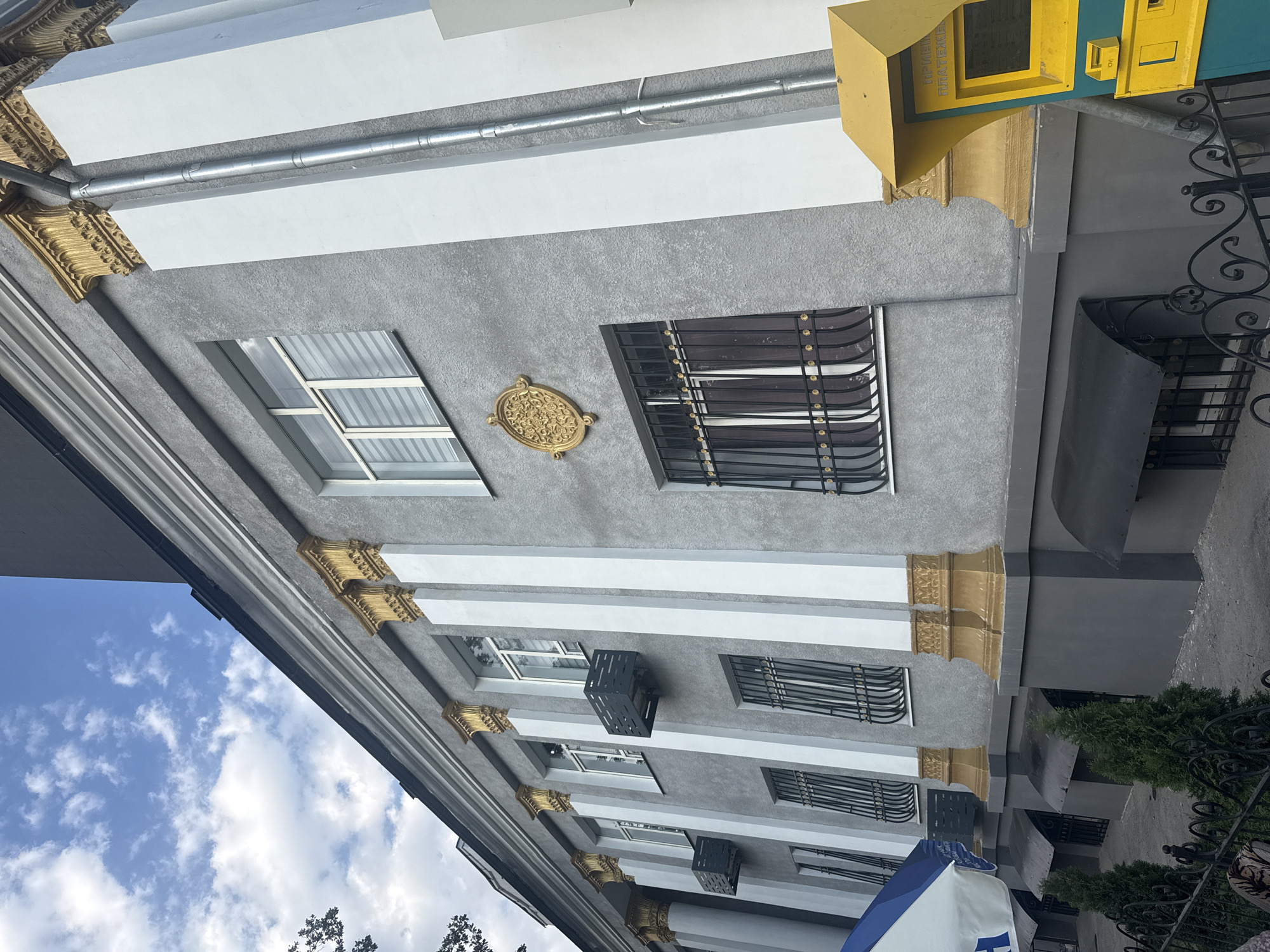 The image shows a portion of a building in Bishkek, Kyrgyzstan. The building has a gray facade with ornate gold columns and white detailing. There are multiple windows, some with decorative black grates. On the ground level, there are awnings over basement windows. A yellow and teal payment kiosk is visible on the right side of the frame. In the foreground, there is a black metal fence and some small evergreen trees. The sky above is blue with scattered white clouds.