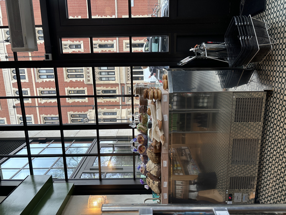 The image presents an interior perspective, likely from a bakery or cafe, looking outward through a large, black-grid window structure onto a street scene in London, United Kingdom. The primary exterior landmark is a prominent, multi-story red brick building directly opposite, identified as "ARUNDEL HOUSE" on its lower facade. This building features numerous Gothic-revival style arched windows with white sills. Visible on the street are parked vehicles, including a dark SUV and a white van. A person with light hair and a dark jacket is discernible near the white van.
In the interior foreground, a display counter occupies the lower center of the frame. It features a light wooden top holding an array of baked goods: various loaves of artisan bread, baguettes wrapped in paper, stacks of bagels, and other pastries. The counter's base is reflective stainless steel with three perforated ventilation grates, mirroring the black and white checkerboard tiled floor. To the far left, a dark green architectural beam and a glowing, woven-texture wall sconce are visible. To the right of the display counter, near a black-framed door, a stack of black wire shopping baskets rests in a metal stand. Adjacent to these baskets is a chrome fire extinguisher. An air conditioning or ventilation unit is mounted above the right window frame.