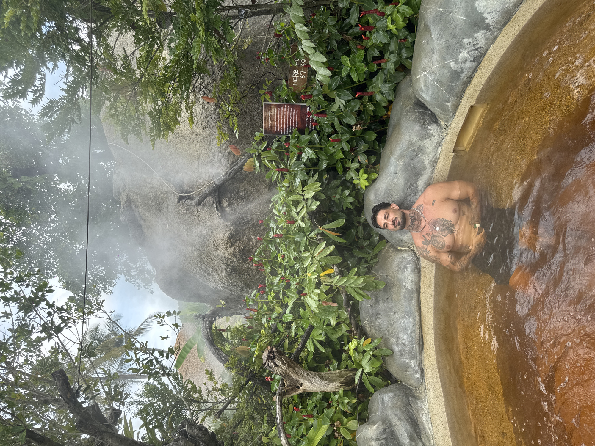 A man with tattoos is sitting in a hot spring, surrounded by lush vegetation. Steam rises from the water and a nearby rock formation. A sign labeled "HERB RO 37-39" is visible amongst the plants. The location is in Ban Khlong Khian, Thailand.