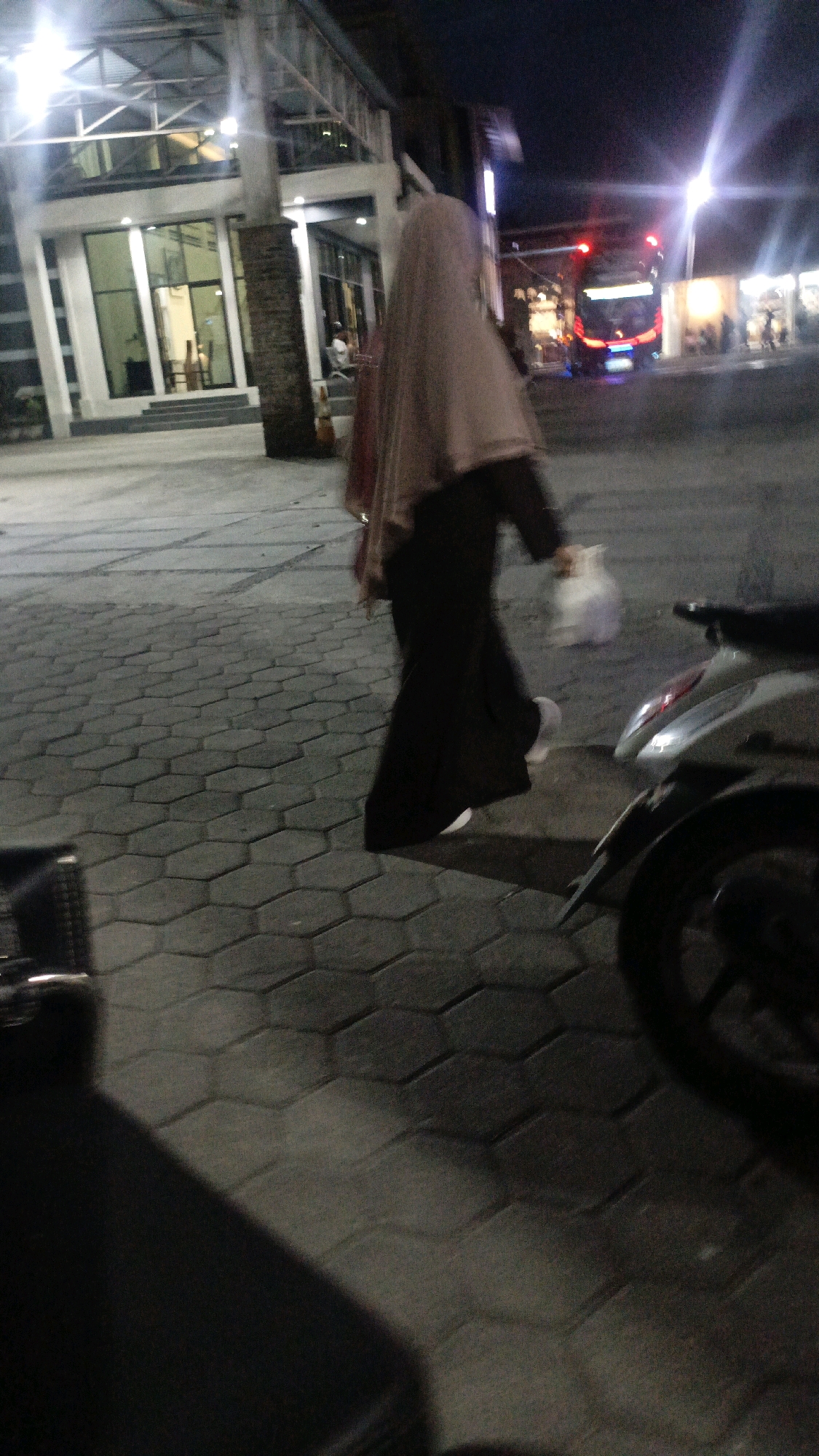 A low-angle, medium shot captures a person wearing a hijab and a dark abaya, walking from left to right across a hexagonal-paved walkway at night. The person is carrying a white plastic bag in their right hand. In the background, a modern building with a glass facade and exposed structural elements is visible, illuminated by interior lights and overhead lamps. To the right of the building, a bus with illuminated red taillights is stopped on a street. A motorcycle is parked in the foreground on the right side of the image, partially obscuring the walkway. The scene is dimly lit, with streetlights and building lights casting shadows on the ground. The image appears to have been taken in the city of Tual, Maluku, Indonesia, likely in a commercial or public area.