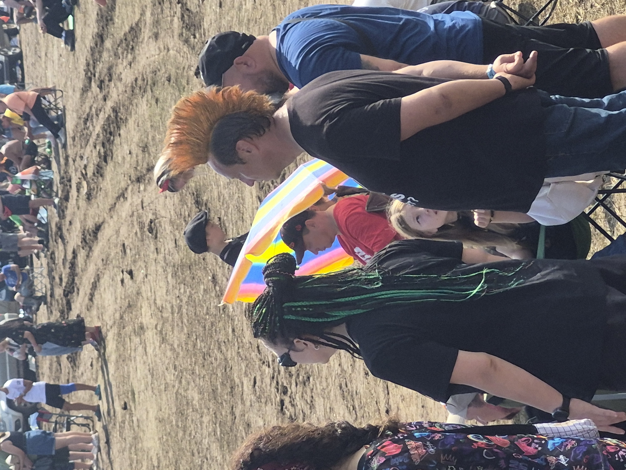 In Lagunovo, Russia, a gathering is taking place outdoors on a dirt ground. The scene is populated by a diverse group of individuals. In the foreground, a person with green-streaked braided hair is wearing sunglasses and a black t-shirt. To their right, a man with a prominent orange mohawk hairstyle is visible, dressed in a black t-shirt and blue jeans. Next to him, another man in a blue t-shirt and black shorts stands, facing towards the right. A colorful umbrella, likely a beach umbrella with yellow, blue, and pink stripes, is partially visible behind the man with the mohawk. In the background, other individuals are scattered around, some sitting in chairs and others standing. The ground appears to be dry and dusty with tire tracks visible. The overall atmosphere suggests an outdoor event or festival.