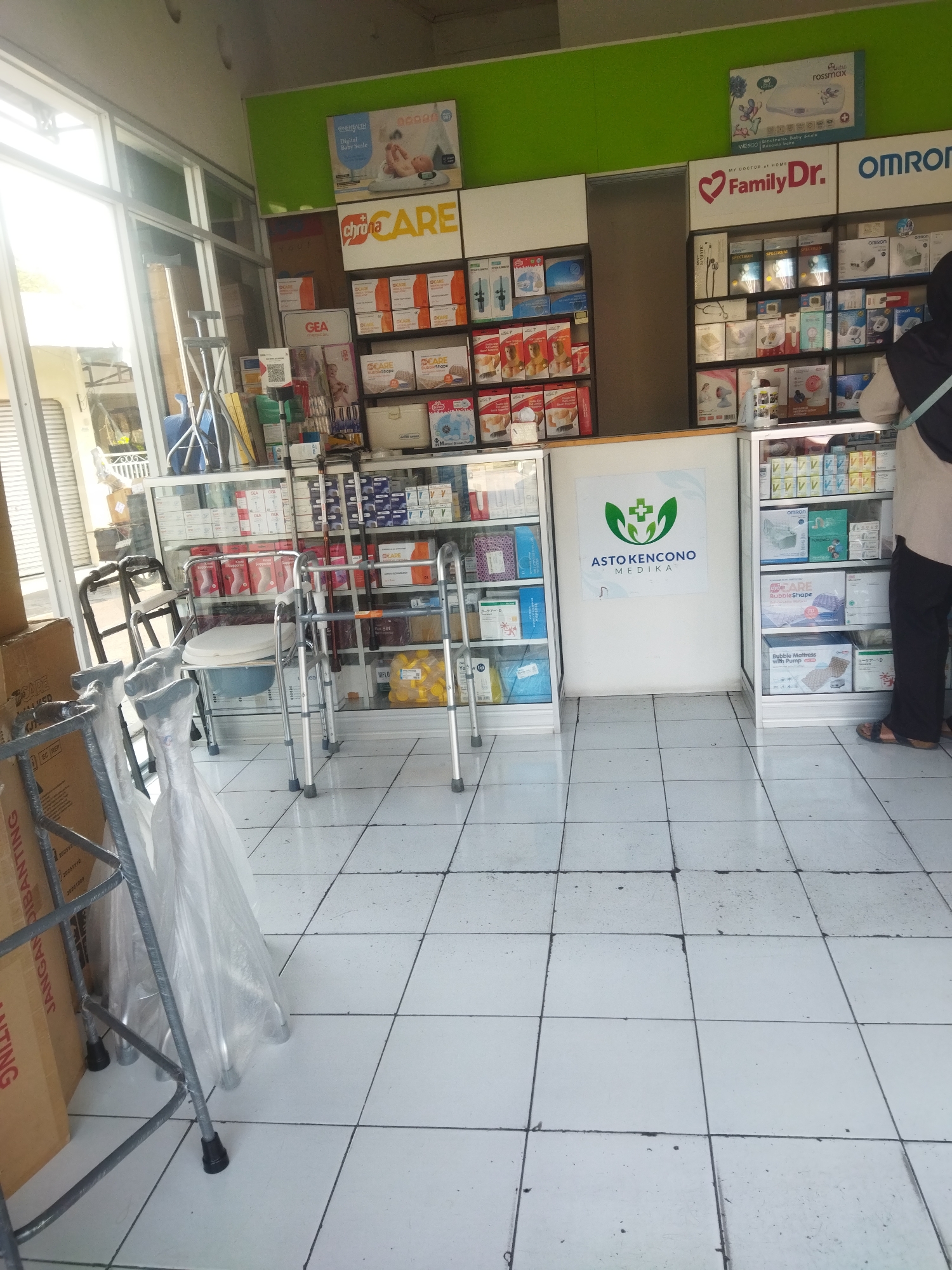 The image shows the interior of a pharmacy, likely "Asto Kencono Medika" based on the logo displayed on the wall. The pharmacy is well-stocked with various medical supplies and pharmaceutical products, displayed on shelves in glass cabinets. Visible items include baby scales, adult diapers, nebulizers, walking aids (walkers and crutches), and various boxes of medication and health products. A person, wearing a dark hijab and a light-colored garment, is standing to the right of the frame, facing away from the camera and towards the shelves. The floor is tiled with white square tiles. The overall impression is of a clean and organized retail space dedicated to health and medical needs. The mention of Sukoharjo, Indonesia, indicates the geographical context of this pharmacy.