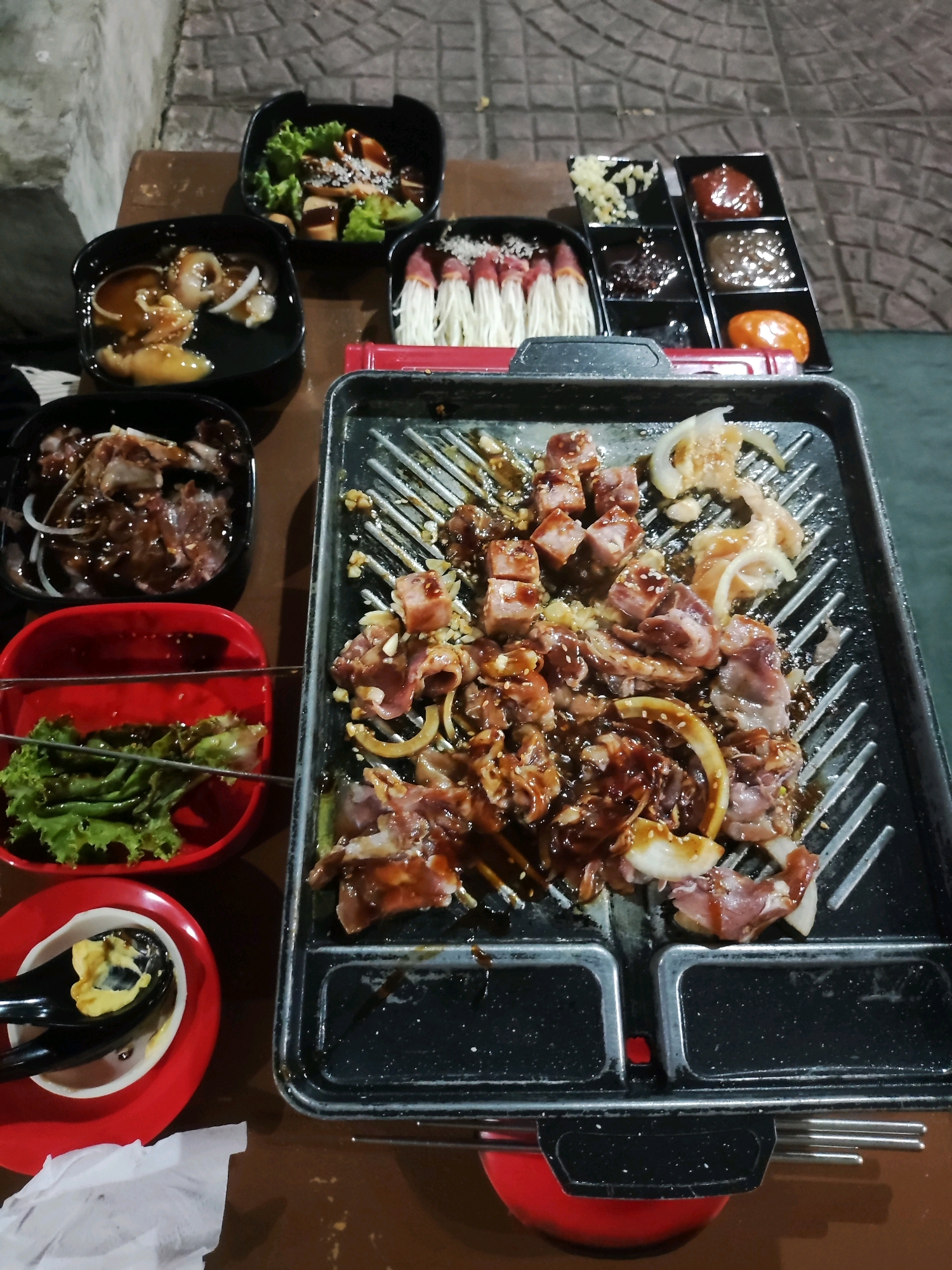 The image depicts an outdoor dining scene, likely at night, in Telagamurni, Indonesia, featuring a tabletop grill laden with marinated meats and onions. The grill is centrally placed on a table, with various small bowls of side dishes and dipping sauces arranged around it. In the background, a stone-paved walkway is visible, suggesting an outdoor restaurant or food stall setting. The lighting appears to be artificial, casting a warm glow on the food. There are no people visible in the image. The scene suggests a meal in progress, with the food ready to be cooked on the grill.
