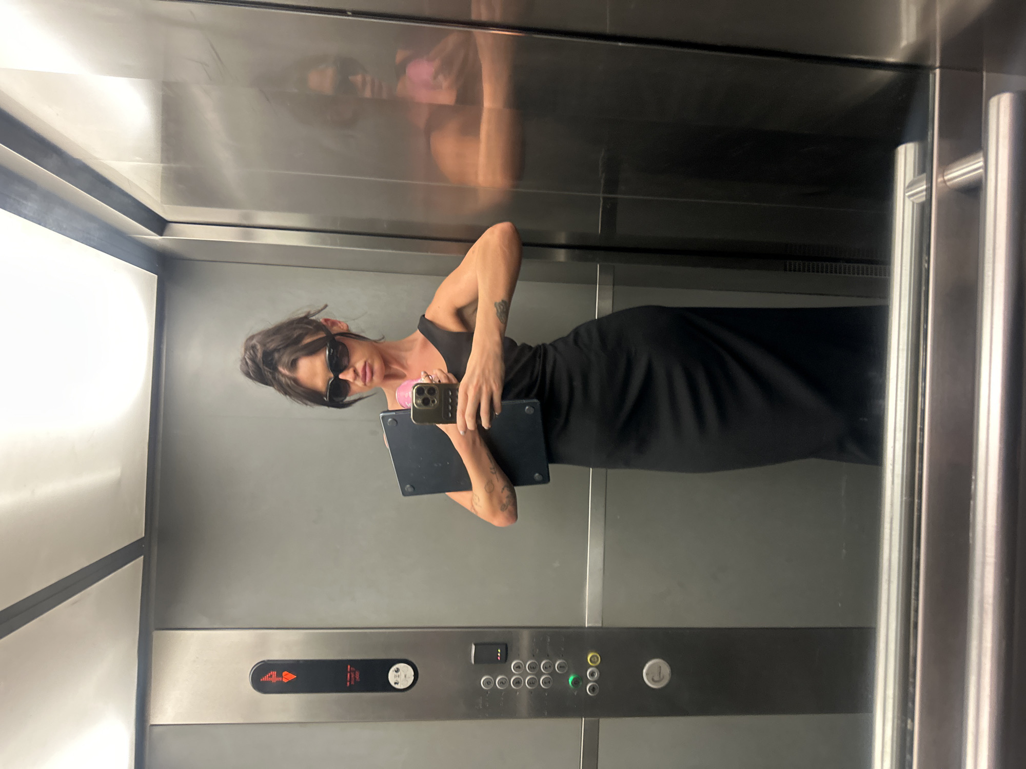 The media file shows a full-length mirror reflection of a person inside an elevator. The person, dressed in a black dress and sunglasses, is holding a laptop and a phone. The elevator appears to be modern, with stainless steel walls and a panel of buttons. One button displays the number "4" in red, indicating the current floor. The reflection in the polished surfaces of the elevator suggests that the image was taken in a well-lit, indoor environment. The overall mood is casual and everyday. There is no visible text in the media file other than the floor number on the elevator panel. The location context of London, United Kingdom, is provided but not discernible from the image itself.