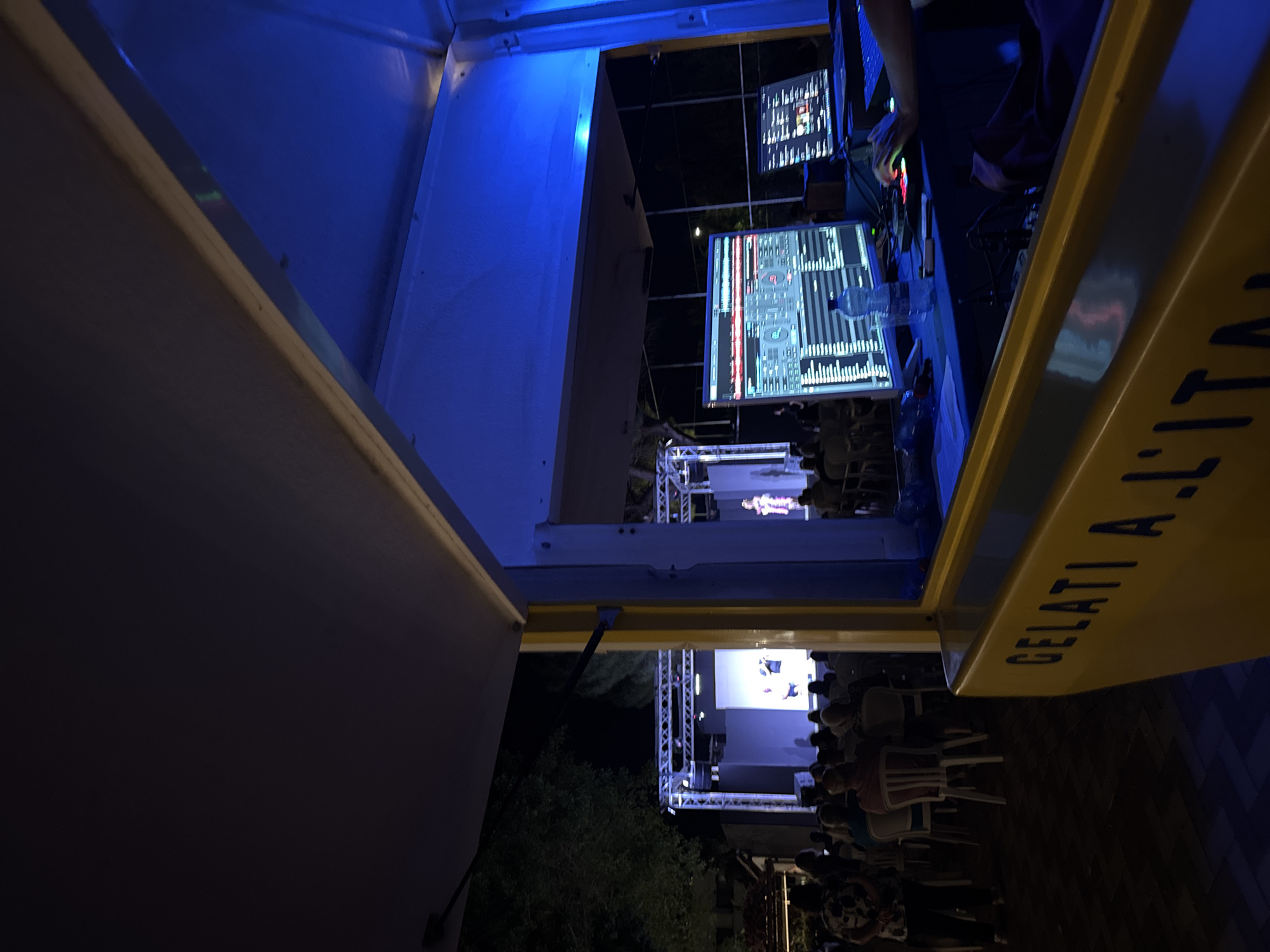 This image depicts an outdoor evening event, possibly a performance or presentation, taking place in Isola delle Femmine, Italy. In the foreground, a yellow structure with "GELATI A L'ITA" inscribed on it is visible, suggesting a gelateria or food stall. Behind this, a DJ setup is present with multiple monitors displaying music software, a hand reaching towards a control deck, and a bottle of water. The background reveals a stage with a large screen, illuminated by blue and white lights, on which performers are visible. An audience is seated in chairs facing the stage, and trees are discernible in the darkness. The overall atmosphere is that of a public gathering during a nighttime event.
