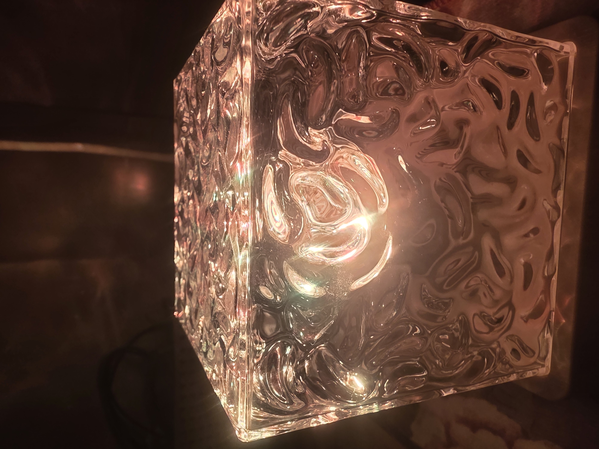A close-up, low-angle shot showcases a clear, textured glass cube with a light source inside. The glass has an organic, wave-like pattern that distorts the light, creating a warm, amber glow. The cube sits on a dark surface, possibly wood, and the background is dark and out of focus, with a hint of what might be a cable or cord on the left side. The scene is dimly lit, creating a moody and atmospheric effect. The image does not depict any specific location in Bila Tserkva, Ukraine, or any people.