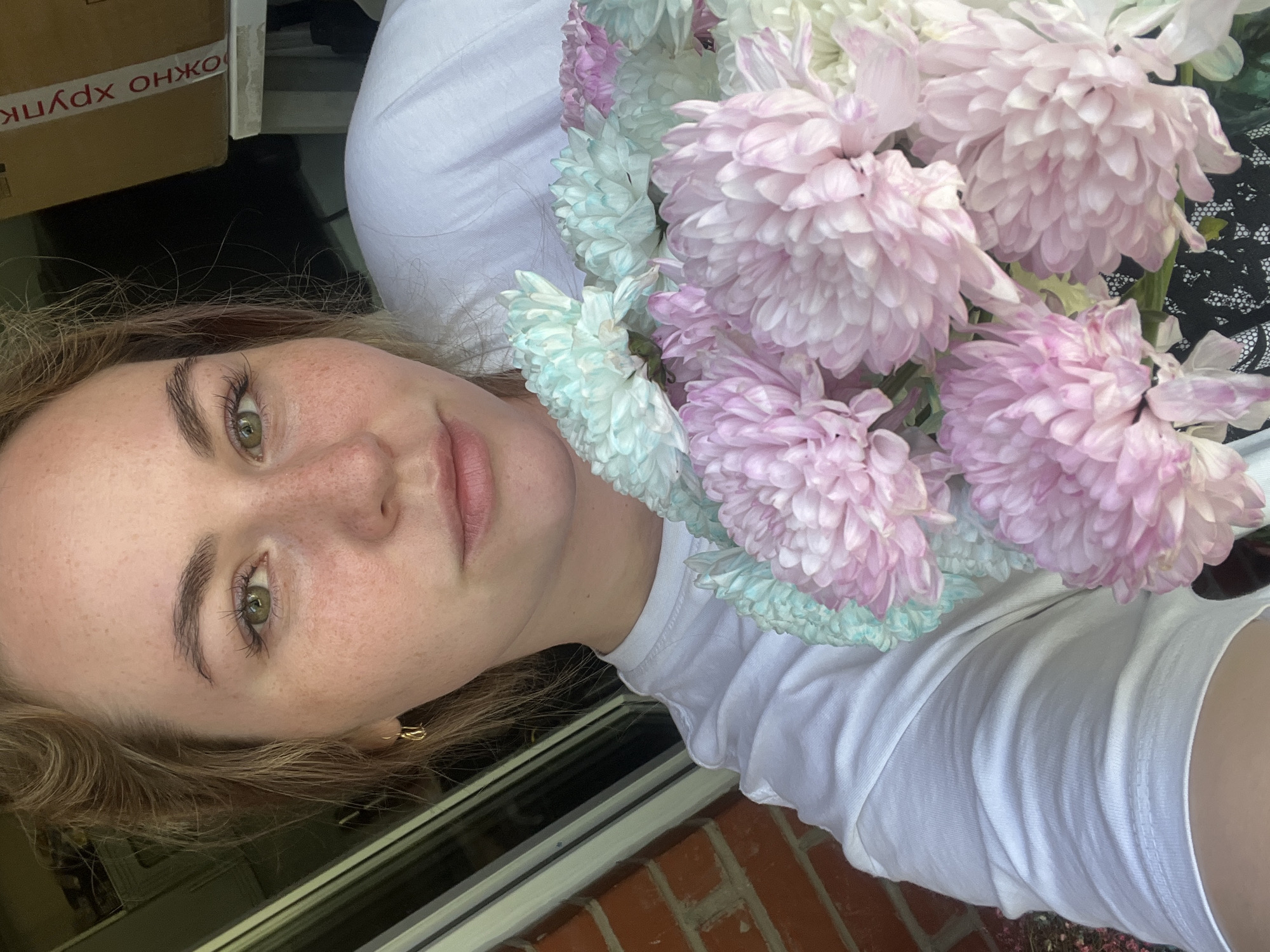 A woman with freckles and green eyes is holding a bouquet of pink and blue chrysanthemums. She is wearing a white t-shirt. The background shows a window frame, a brick wall, and a cardboard box with Russian text. It's impossible to confirm the location as Chaikovsky, Russia, without additional information.