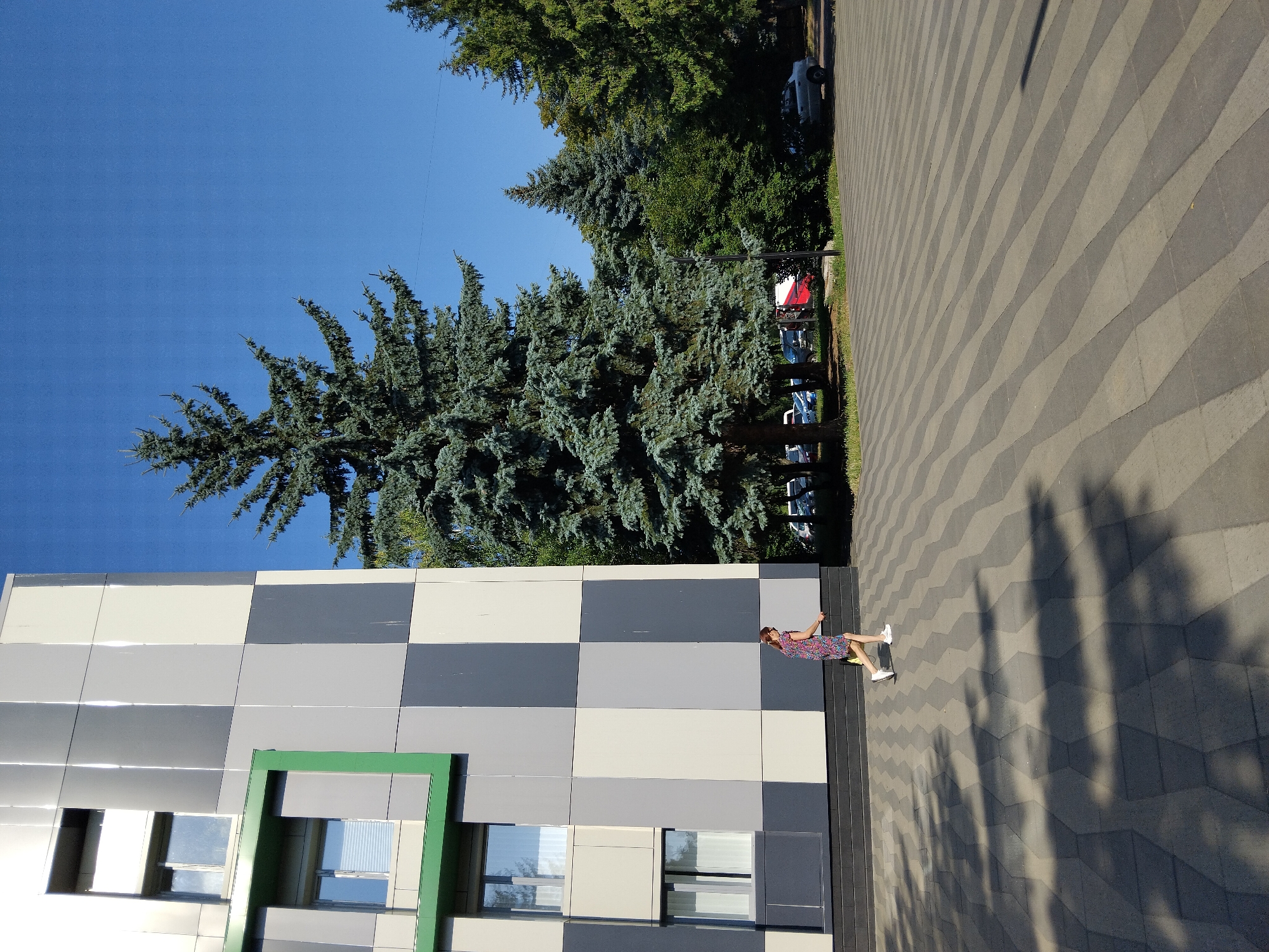 The image shows a person in a colorful dress walking on a patterned paved area in front of a modern building with a grey and white checkered facade. Tall evergreen trees are visible behind the building, and parked cars are seen in the background. The sky is clear and blue. The location is Zhytomyr, Ukraine.